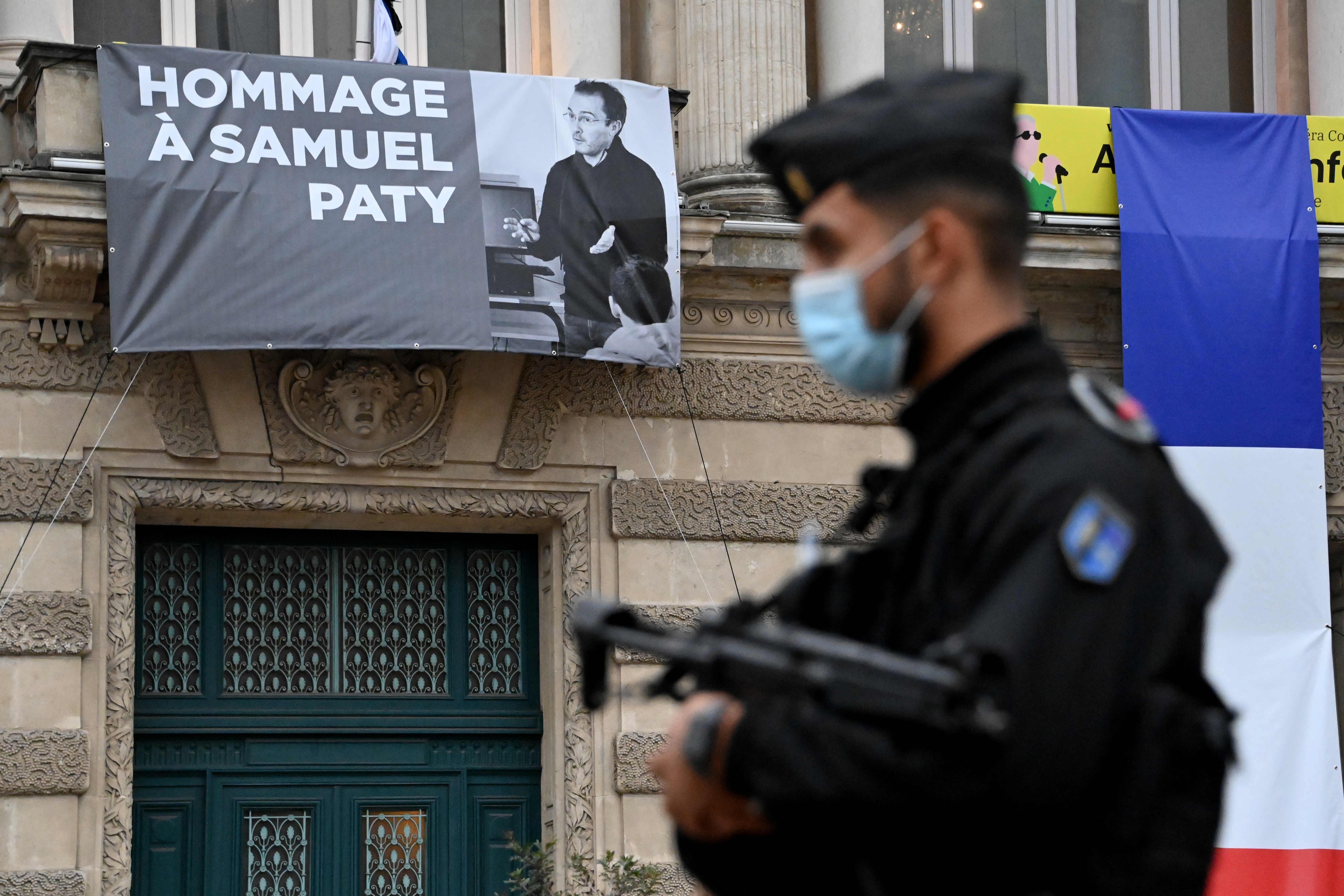 A French police officer stands next to a portrait of French teacher Samuel Paty on display on the facade of the Opera Comedie in Montpellier on October 21, 2020, during a national homage [Pascal Guyot/AFP]