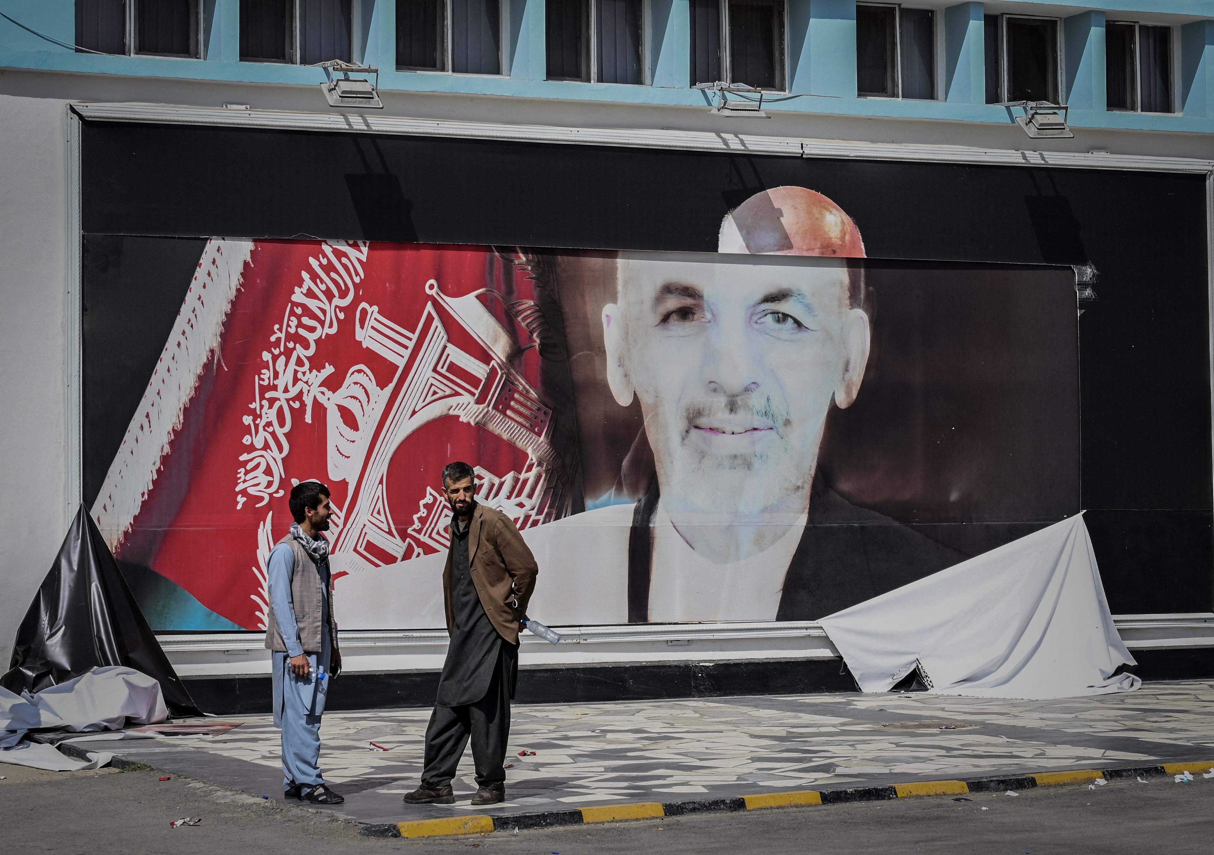 Afghan men stand next to a torn poster of former Afghan President Ashraf Ghani