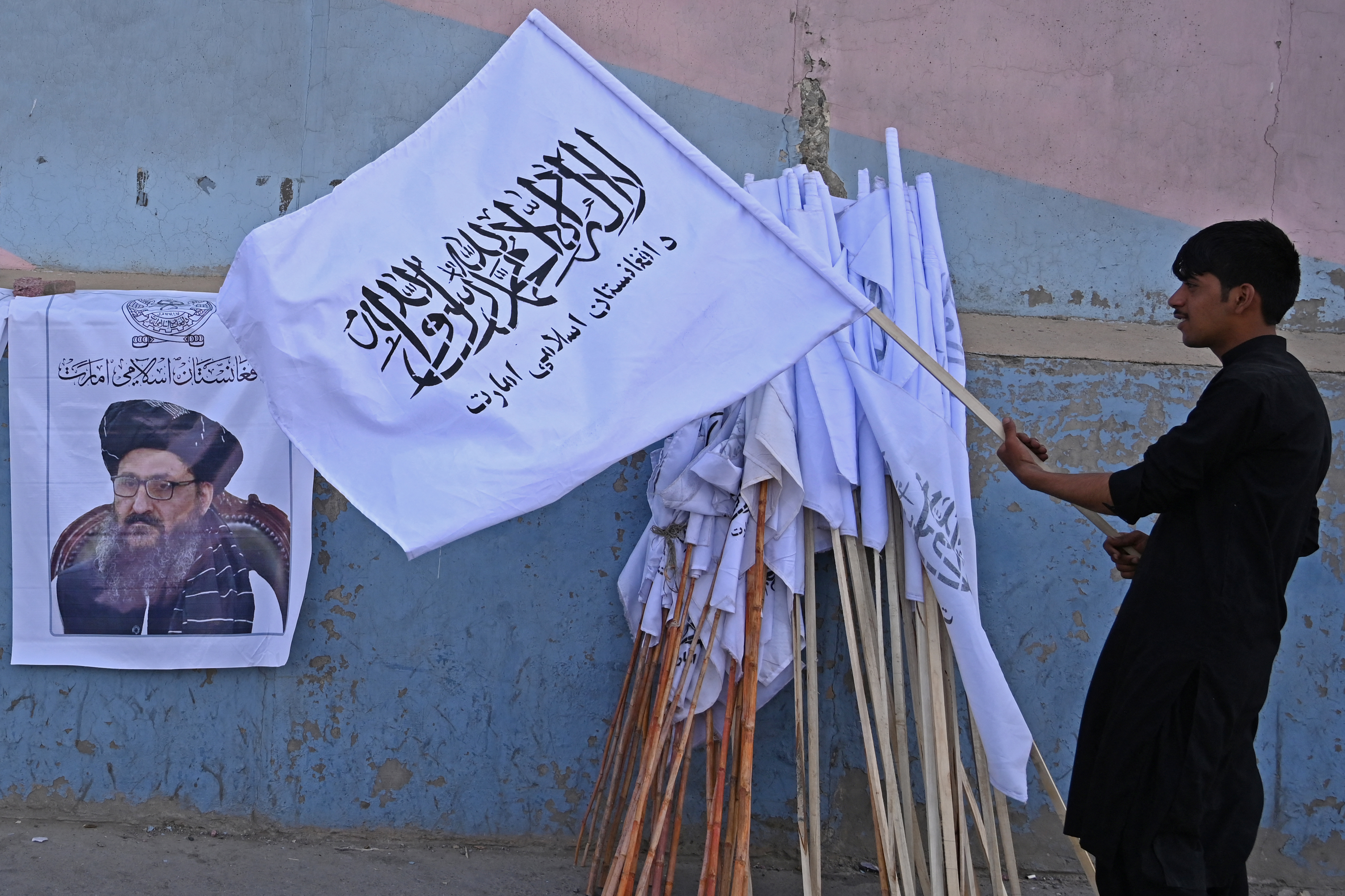 A vendor holds a Taliban flag next to a poster of Taliban leader Abdul Ghani Baradar as he waits for customers along a street in Kabul on August 27, 2021 [Aamir Qureshi/ AFP]