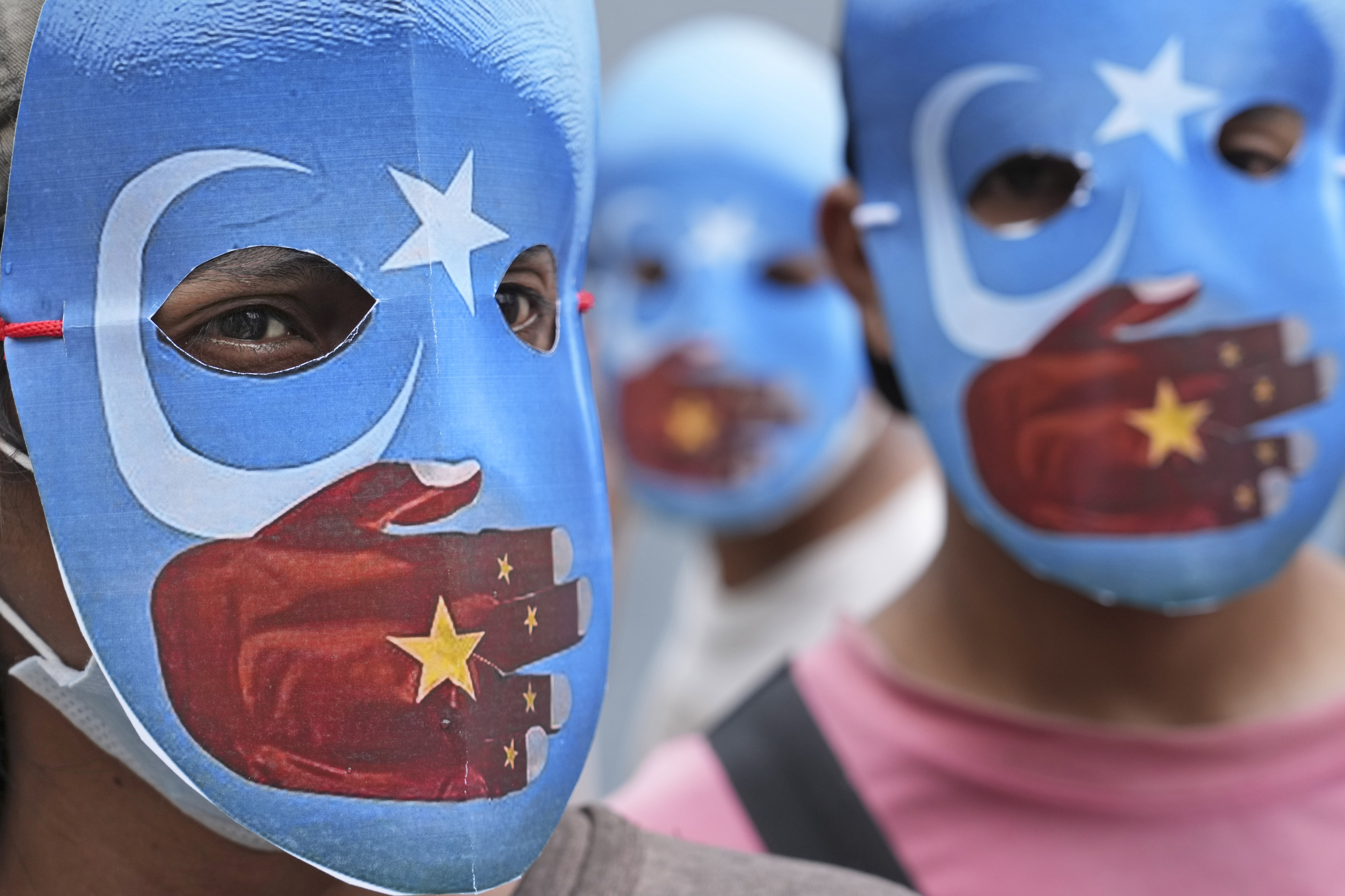 A close up of the faces of three students wearing masks in the colour of pro-independence East Turkistan flag with a hand in the colour of a Chinese flag over their mouths