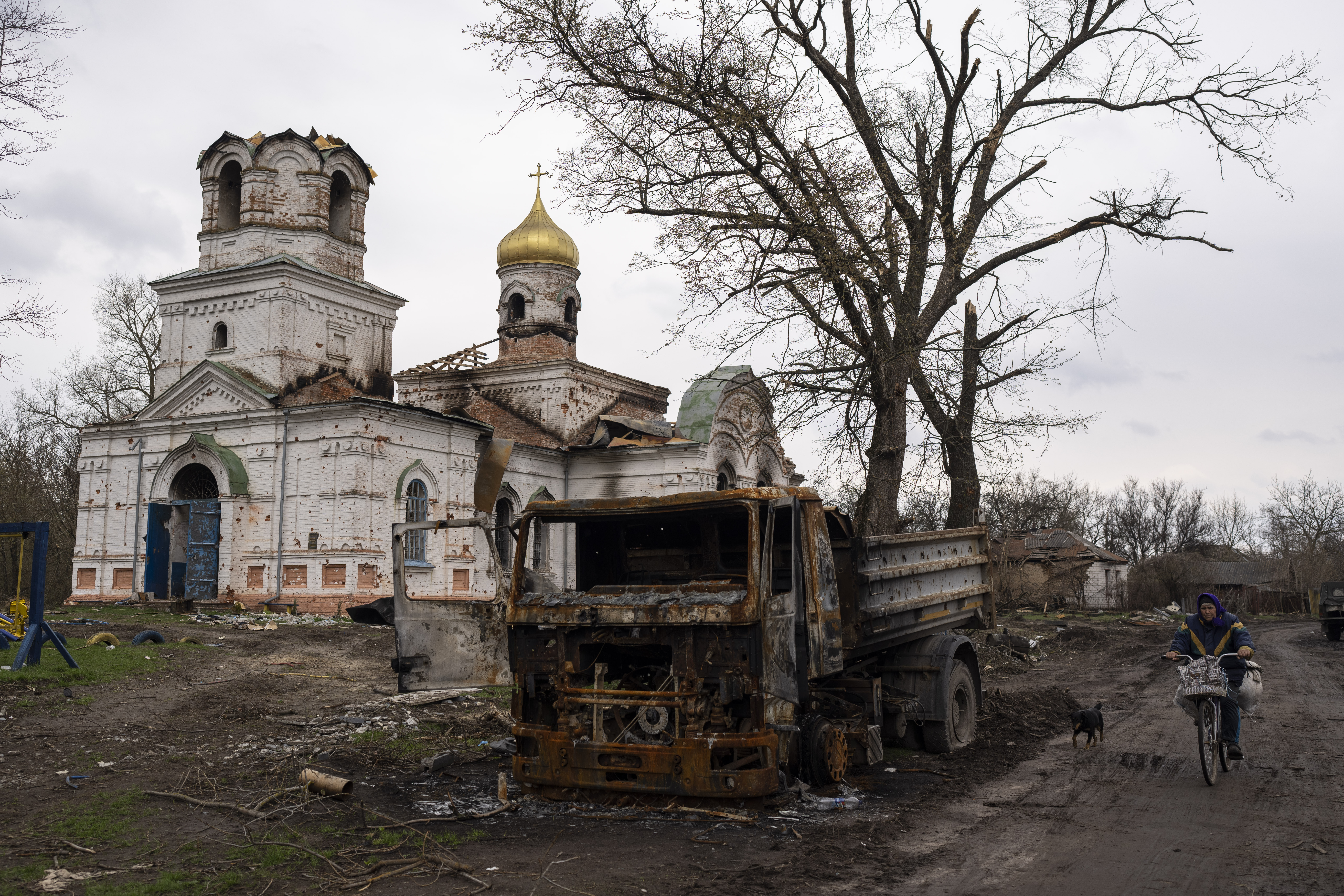 A woman with a bicycle rides by a burned vehicle, in front of a damaged church