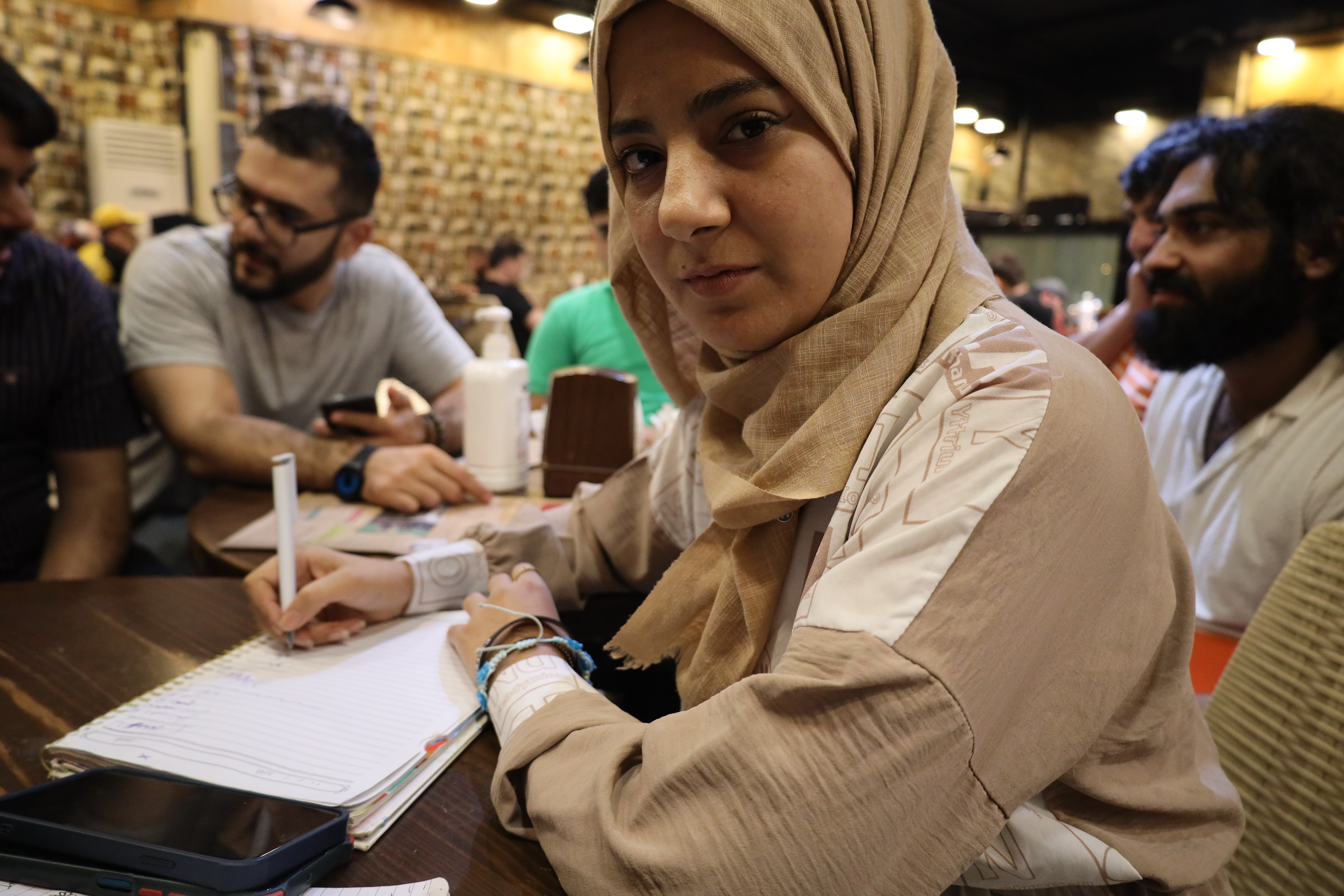 A young woman looks at the camera while writing in a notebook. She is sitting at a table in a cafe with three male friends.