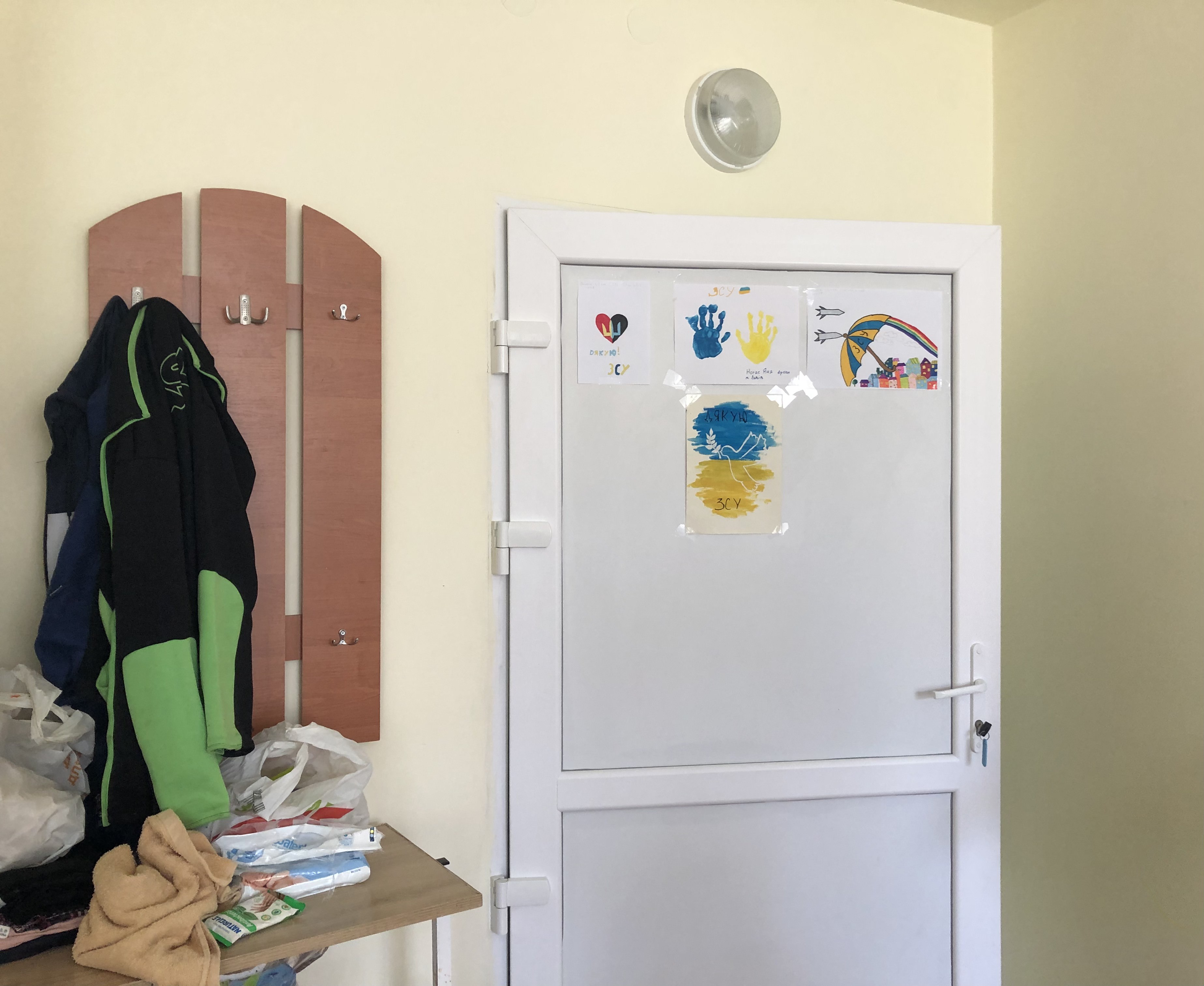 A photo of a door to Pelipenko's room with four drawings/paintings on it, one with a blue and yellow hand print, one with a white crane carved between a drawing with yellow and blue paint, the third painting/drawing is of a large group of people holding an umbrellla and a rainbow in the back and the fourth drawing is a little drawing of a colourful heart.