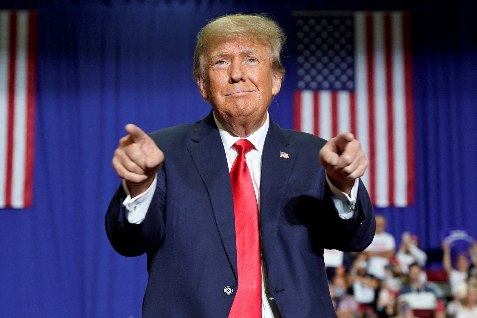 Donald Trump in suit and red tie, pointing with both hands with US flag behind him
