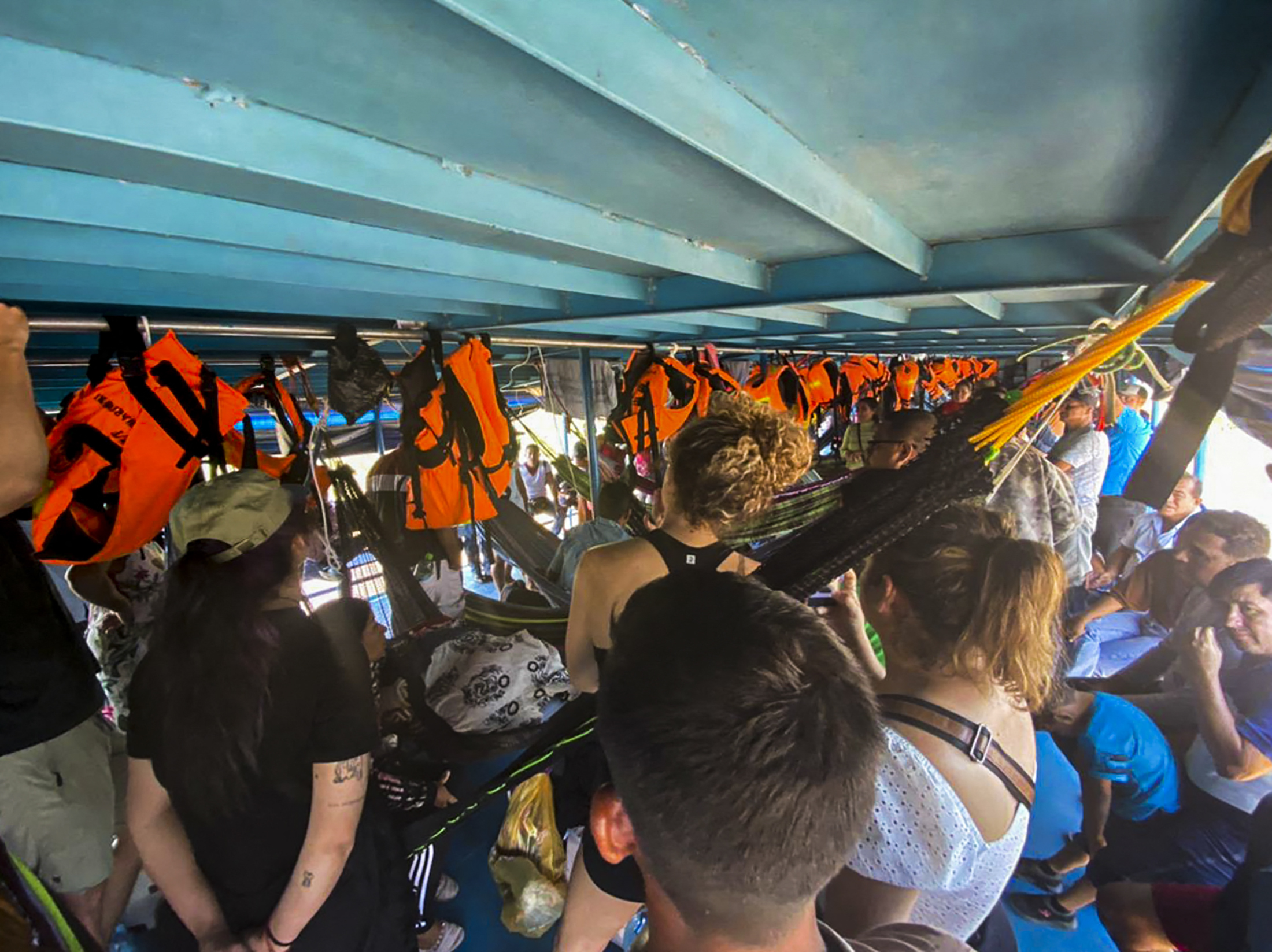 Tourists crammed into a low-ceilinged boat, some standing some sitting, while orange life jackets hang along the side