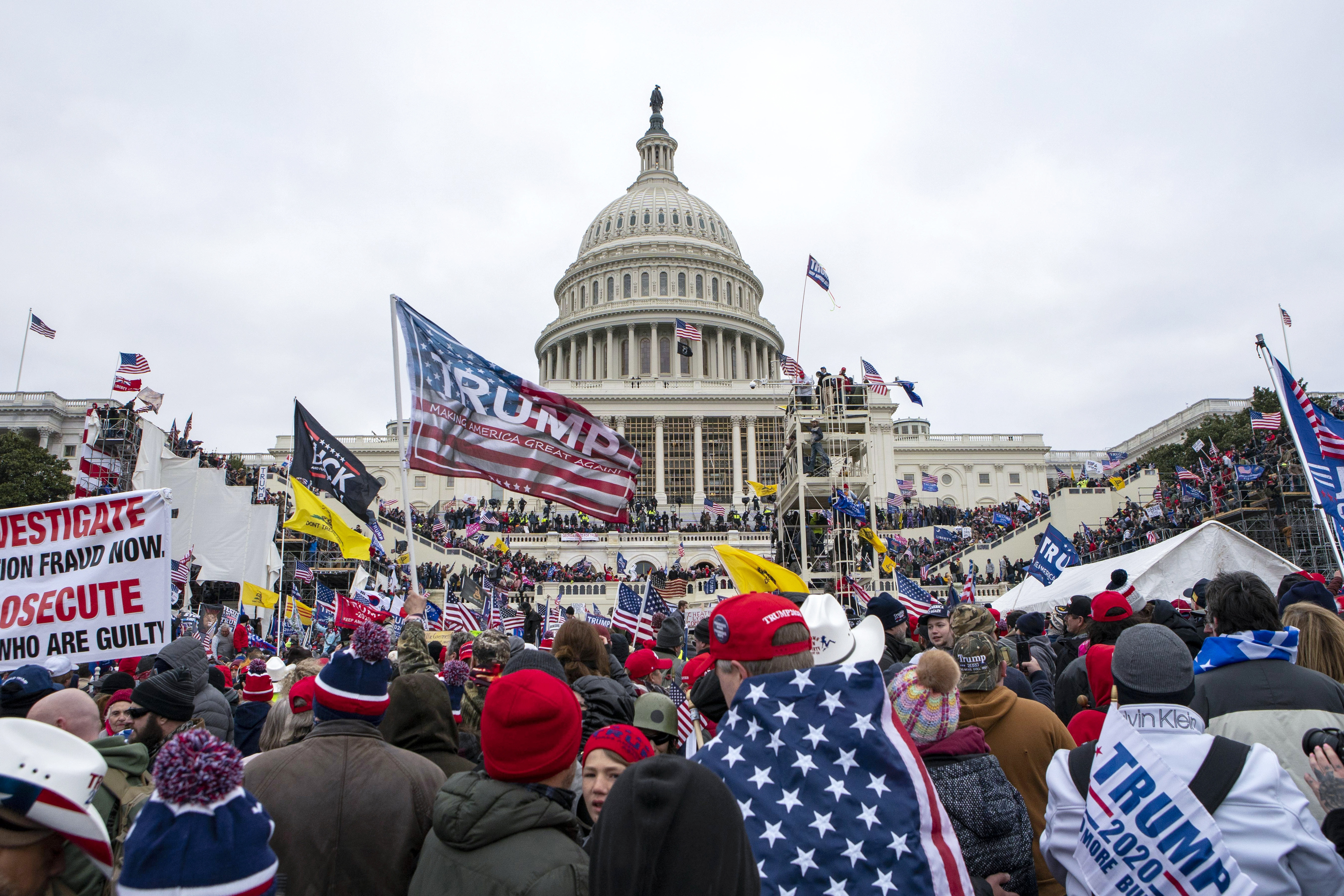 Rioters in front of Capitol building, some with the US flag draped over them, others holding banners that read "Trump". Many are holding the US flag.