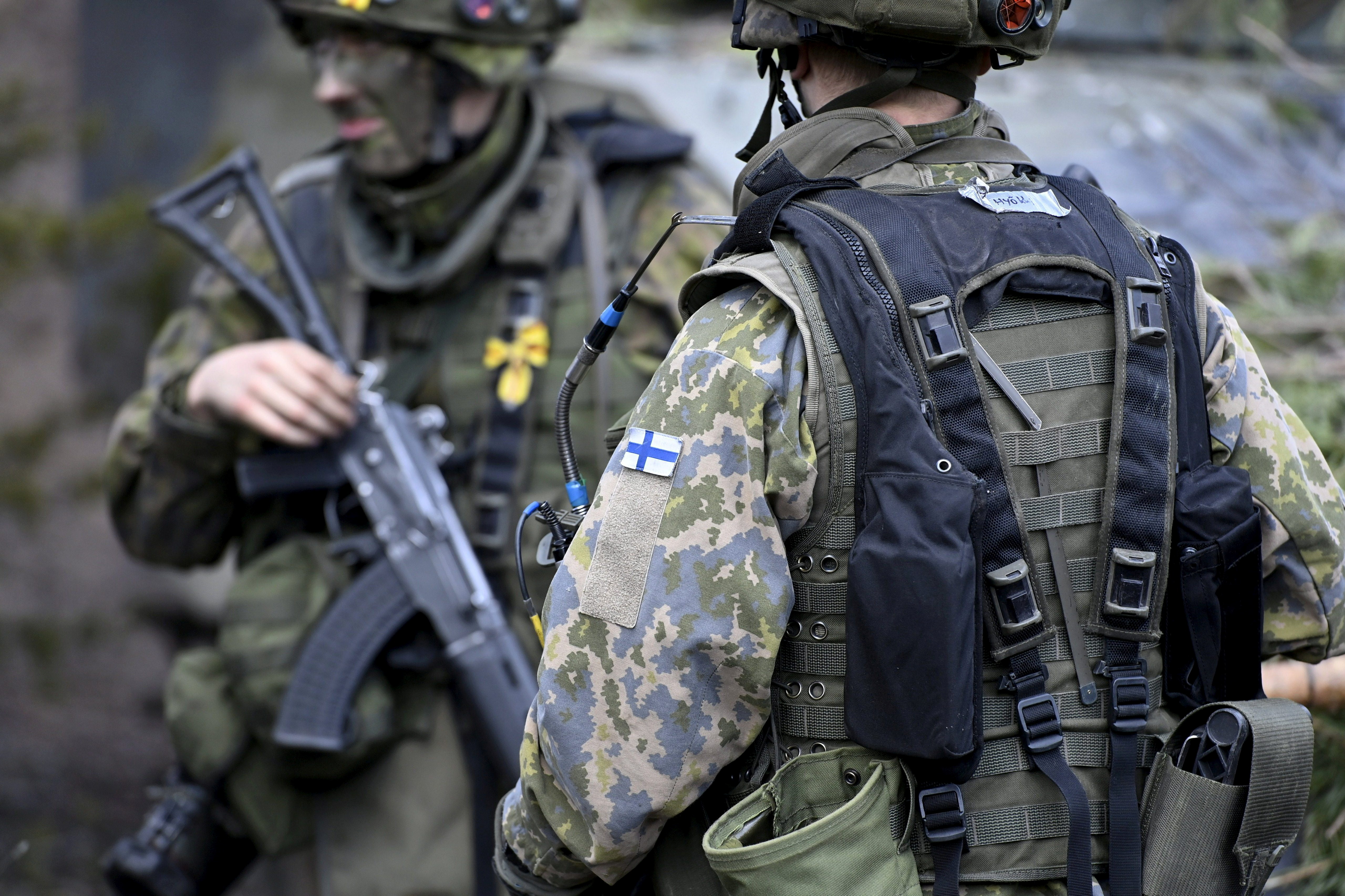 Two Finnish soldiers in combat uniform. One has his back to the camera with the Finnish flag visible on his left shoulder. The other is holding his weapon and side-on to the camera.