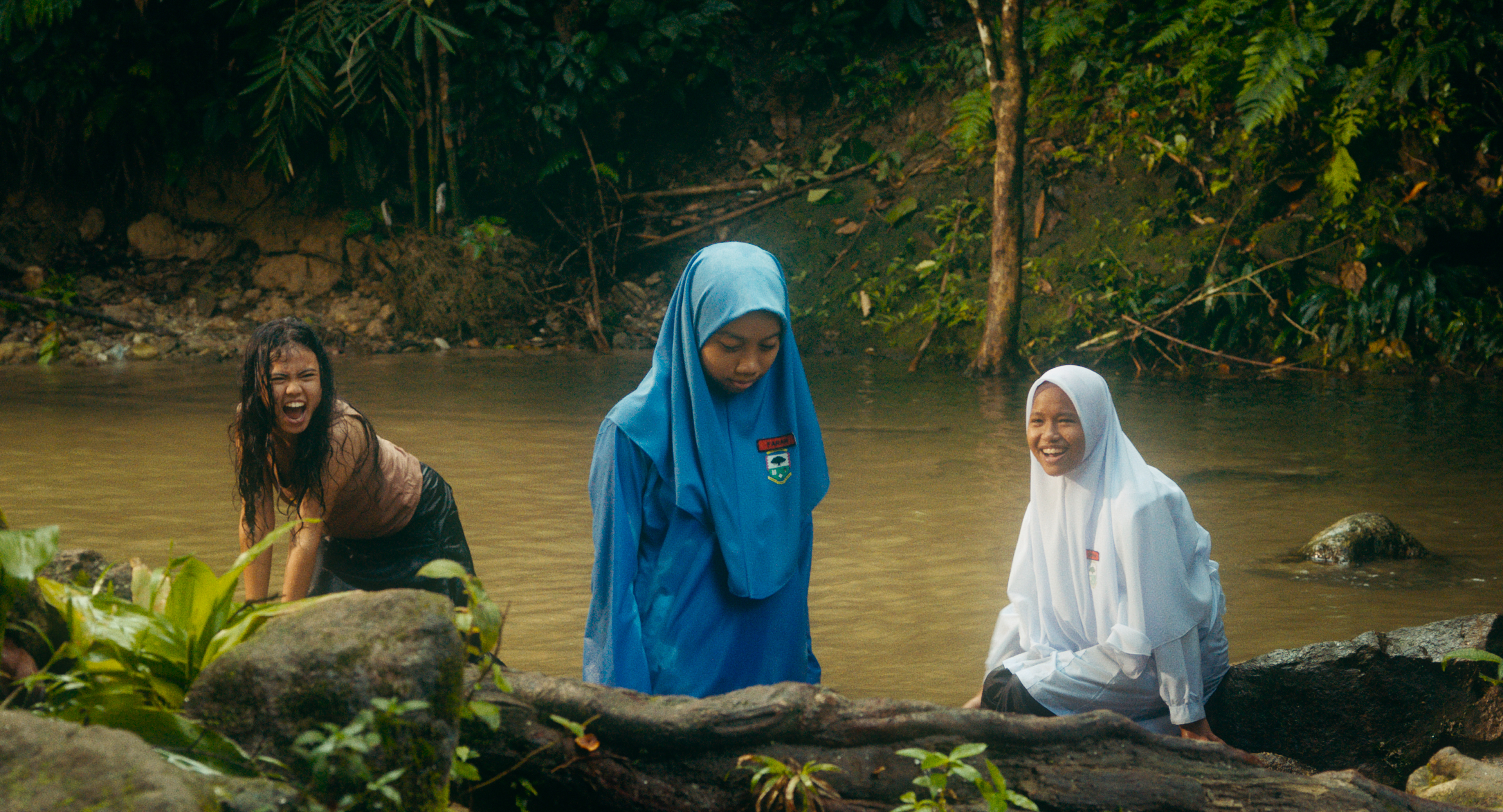 A still from Tiger Stripes showing three girls standing in a river in Malaysia. Two are in school uniform and wearing head scarves. The third is behind them crouching down. She's wearing a black t-shirt and her hair is loose and messy. The river water is brown ad there are trees behind.