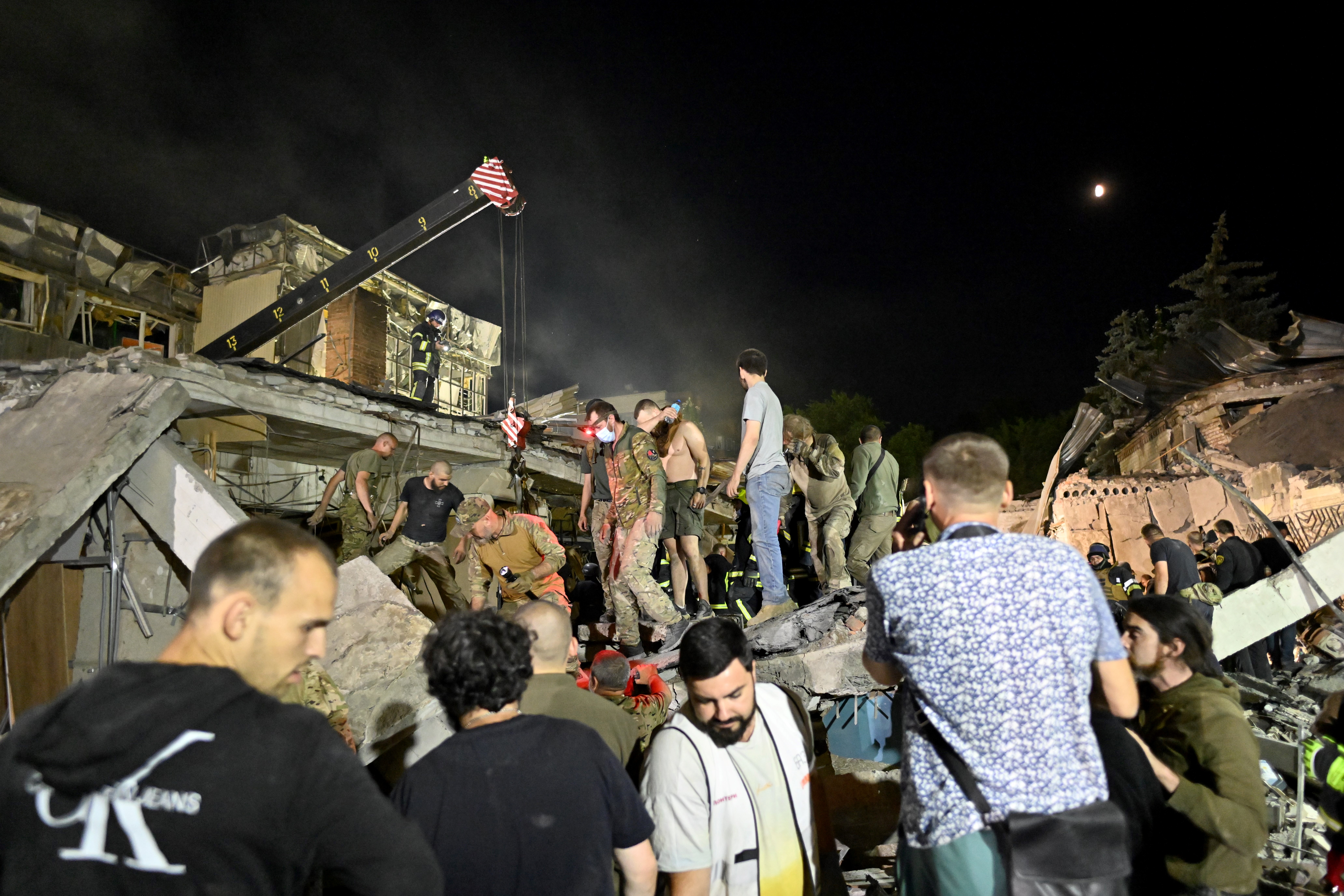 Rescuers and volunteers work with cranes to free people after a Russian missile attack that hit a pizza restaurant. The building has been reduced to rubble. The sky is dark.