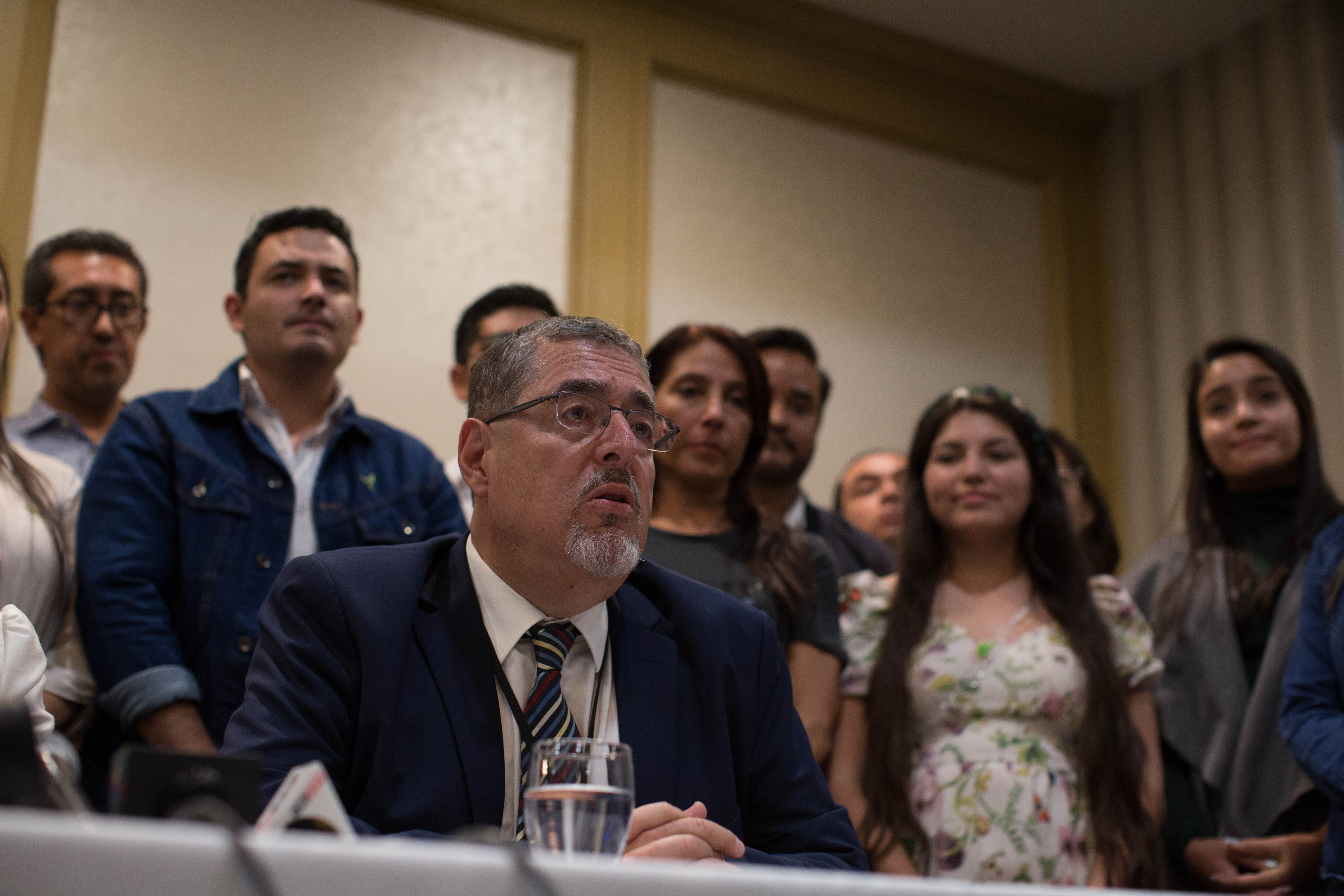 A man in a suit and tie sits at a table and speaks, while supporters line up behind him.