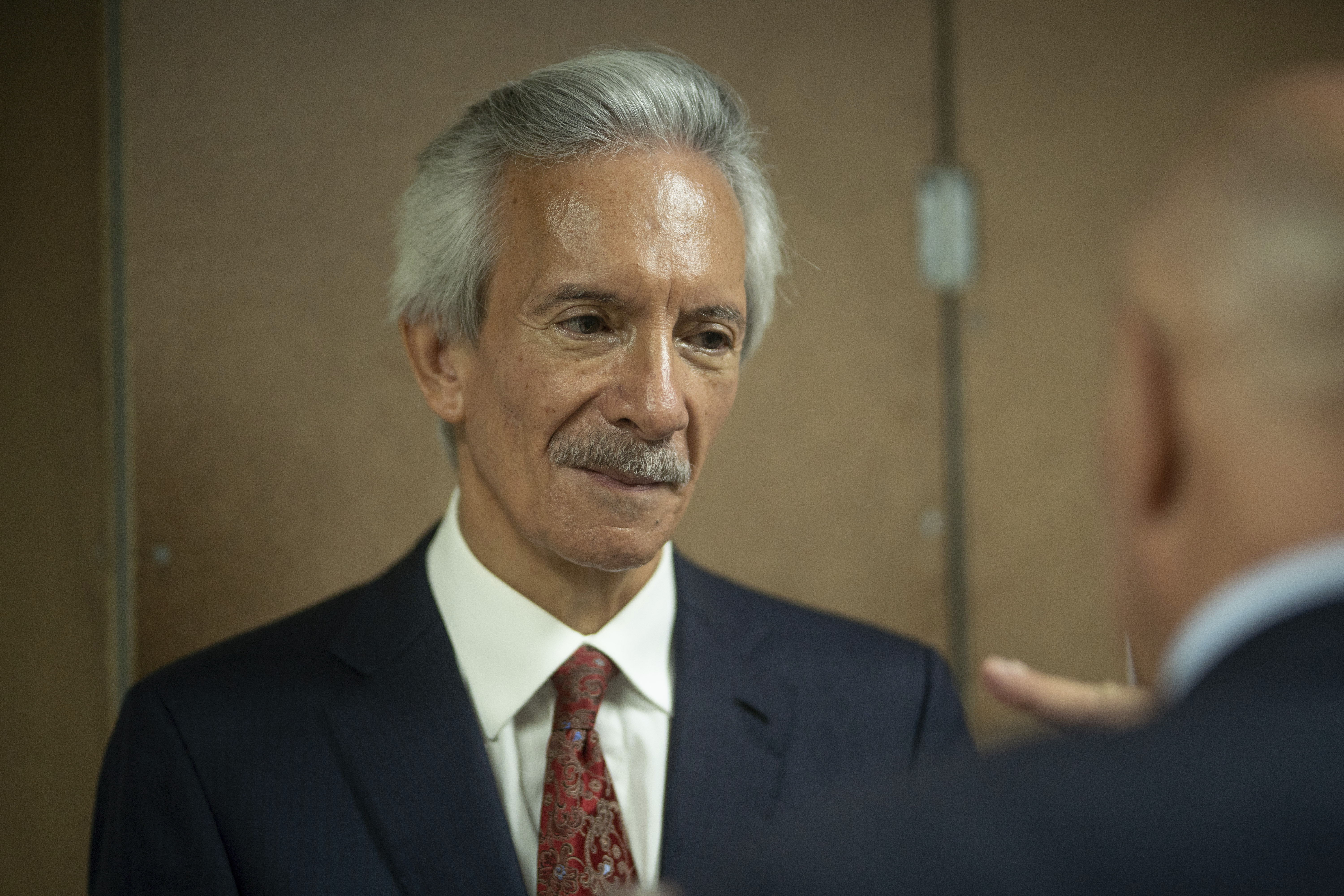 A close-up of an older man with a mustache in a dark suit and red tie.