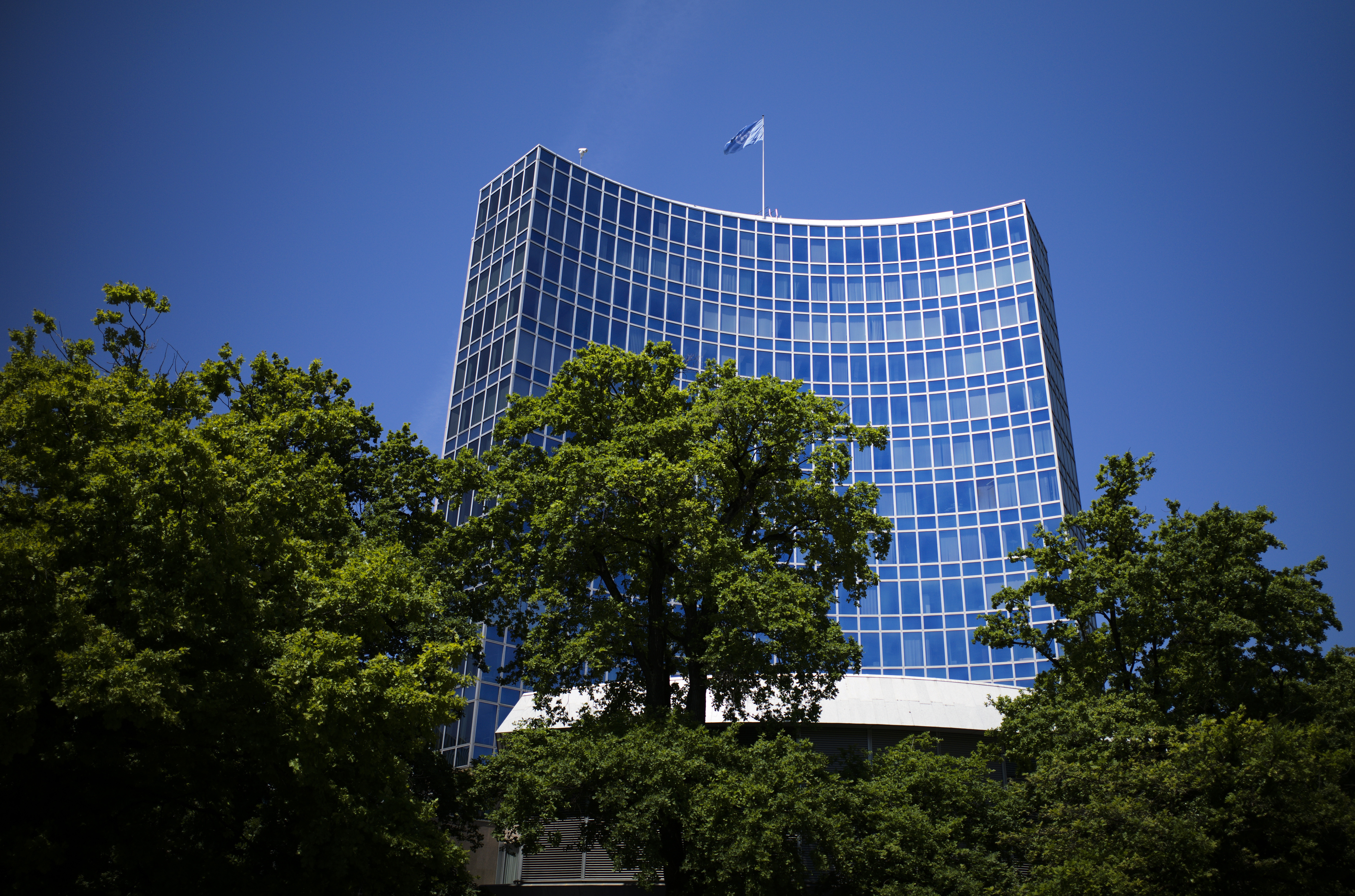 A view of the UN building in Geneva. The sky is blue. There are trees around it, and a UN flag flying from the roof