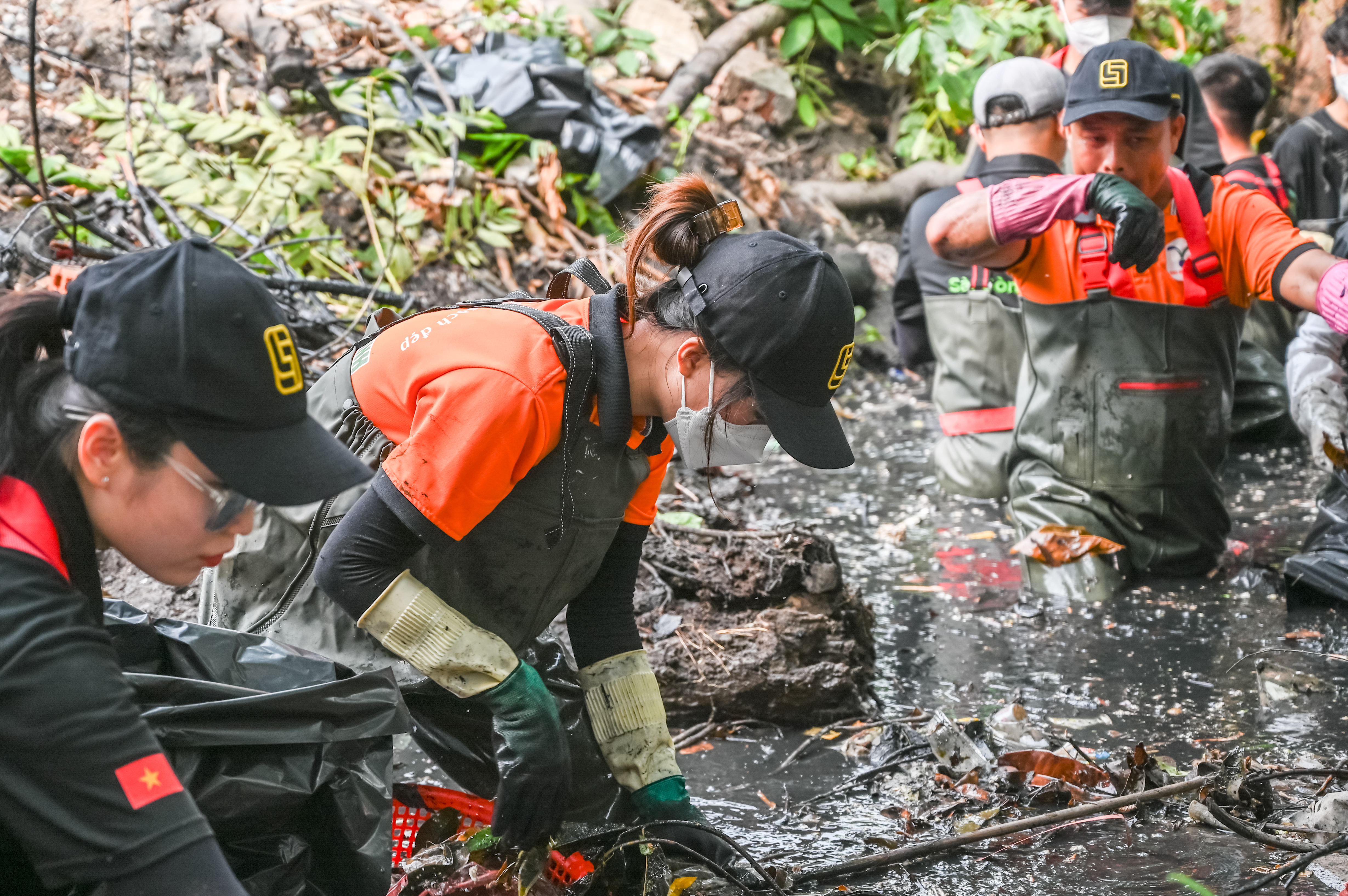 Young people cleaning up the putrid canal. The water is black and filled with rubbish. The cleaners are wearing rubber suits and gloves,