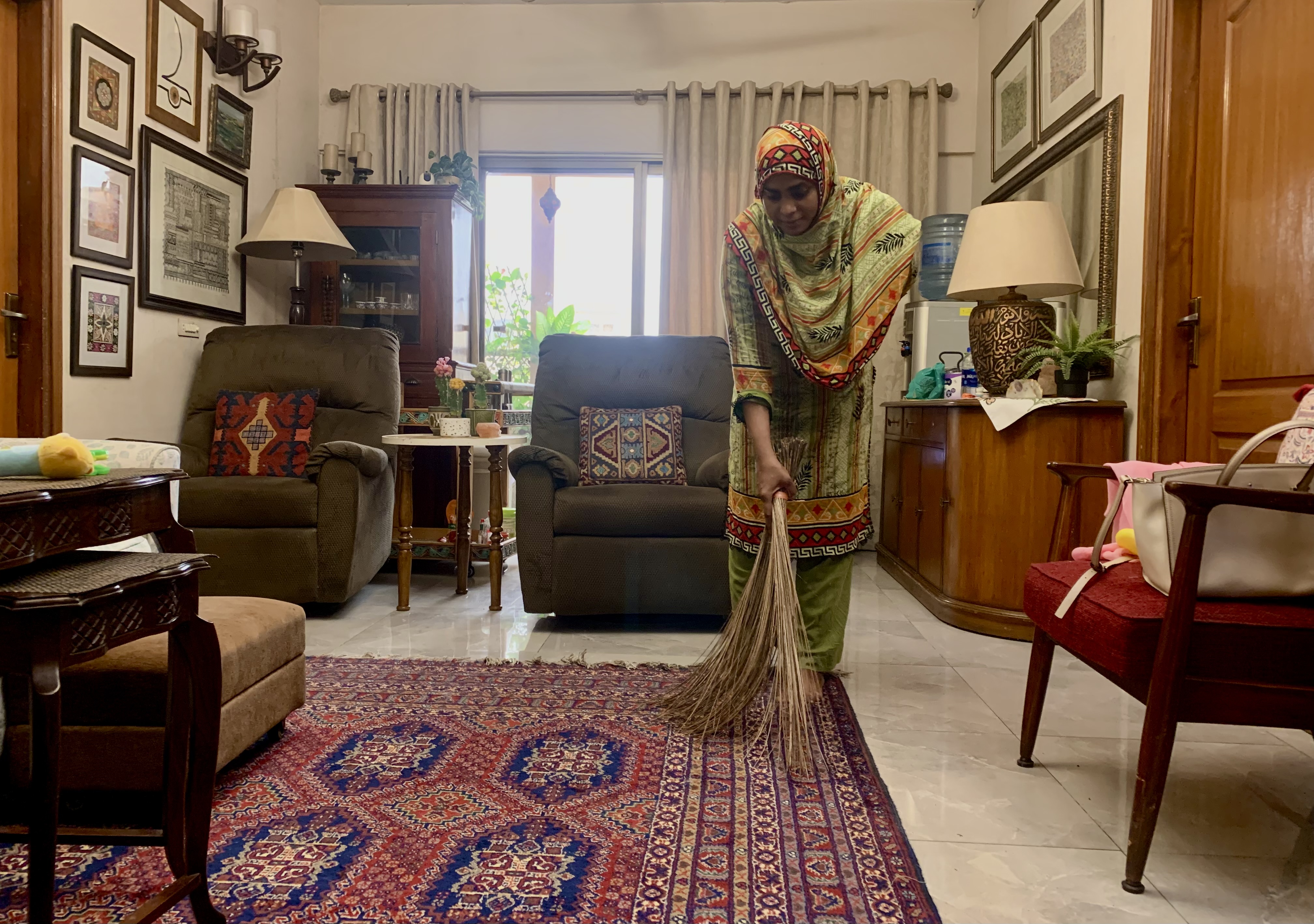 A photo of Aisha cleaning a carpet using a broom with chairs around her and tables and shelves on the walls around her.