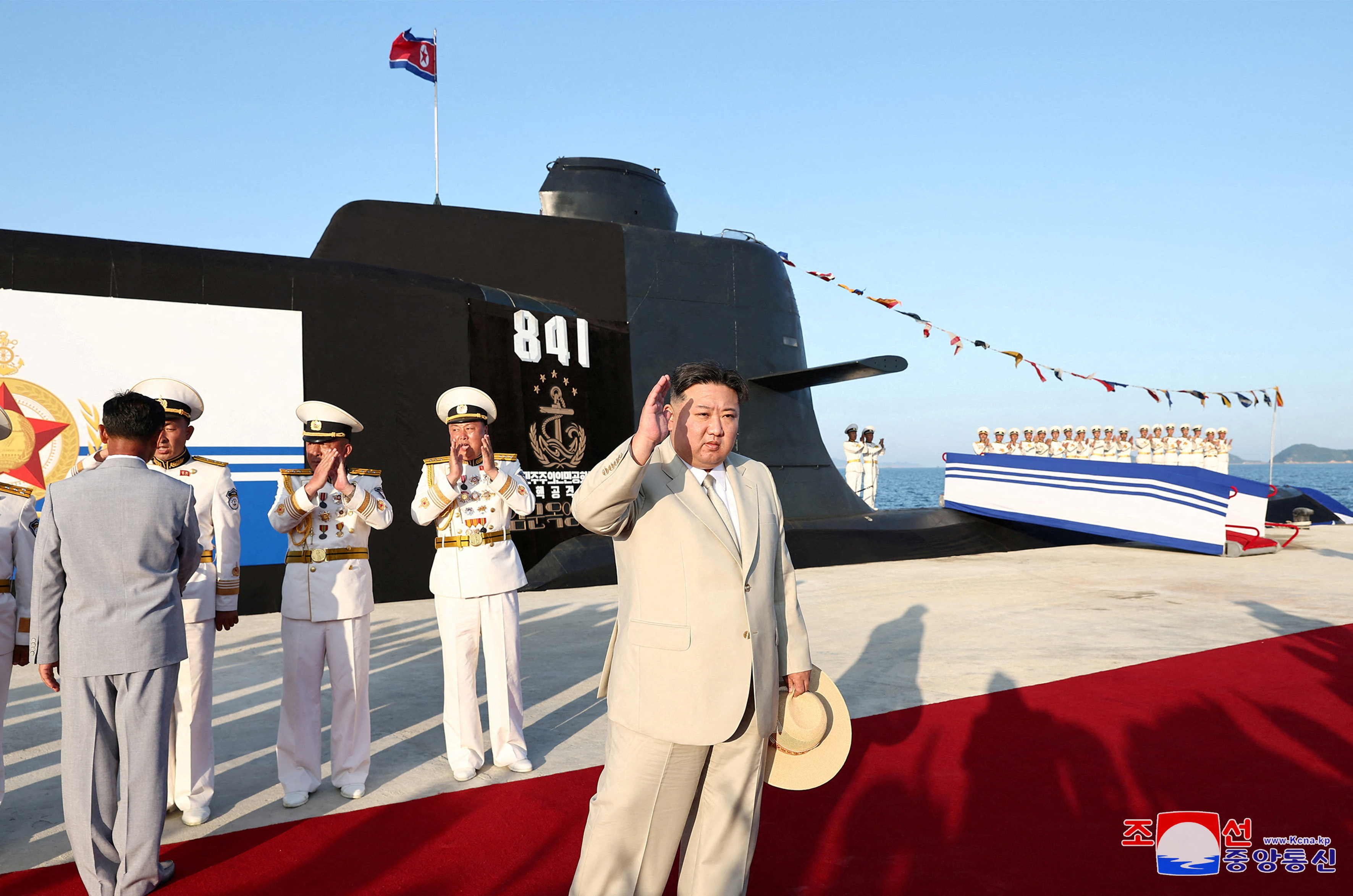 Kim waves at the launch of a new submarine. He is wearing a pale linen suit and carrying a straw hat. The submarine is behind him, It is black with the number 841 written on the side.