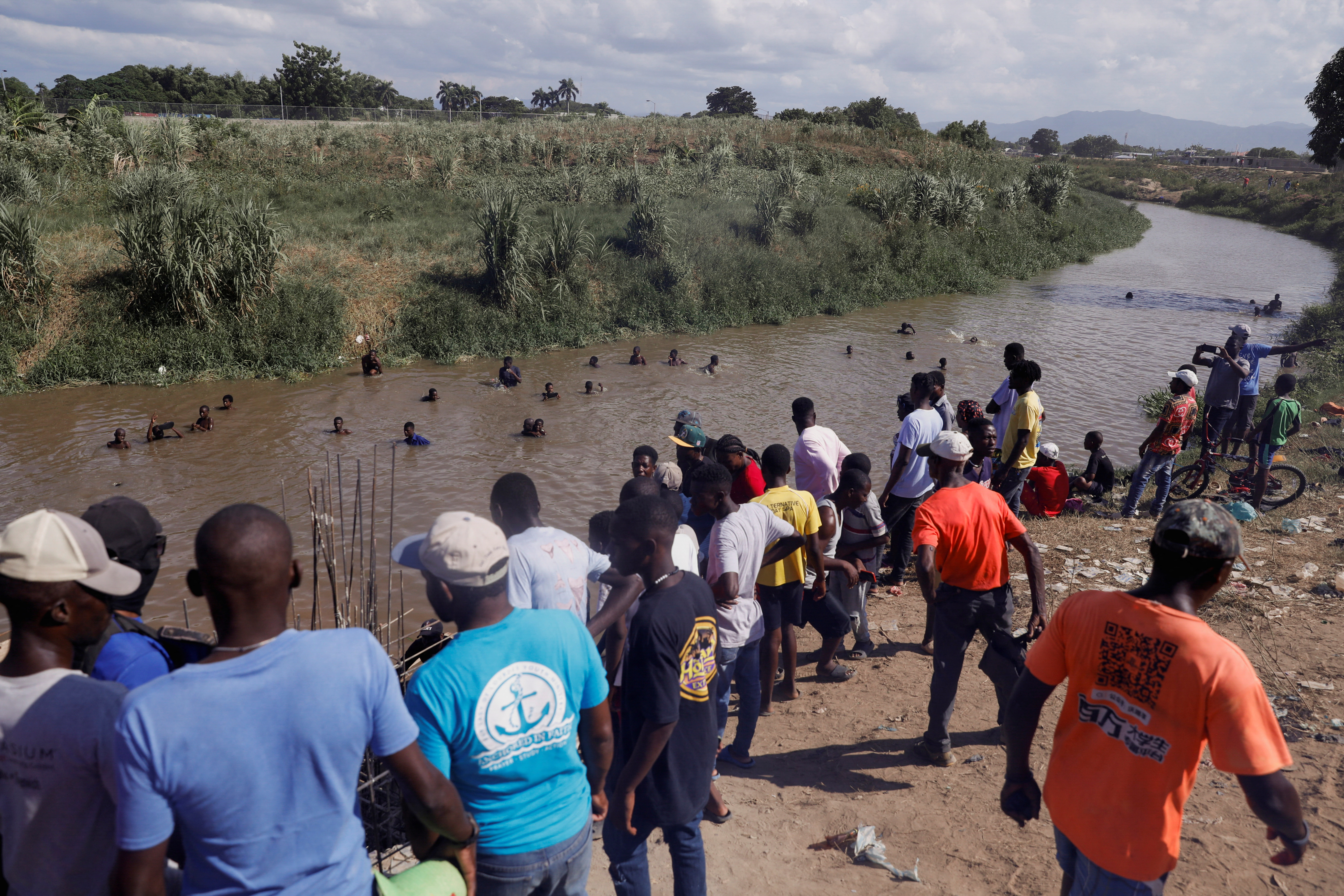 Haitians stand near the constructions site of a water channel of the Massacre river, a river shared between Haiti and the Dominican Republic, after Dominican President Luis Abinader announced a total border shutdown amid a conflict over the construction of the channel, in Ouanaminthe, Haiti September 15, 2023. Many of the people are gathered on the banks of the river, which is covered in brown dirt, while others swim in the water itself, their heads bobbing above the surface. On the other side, a green river bank can be seen.