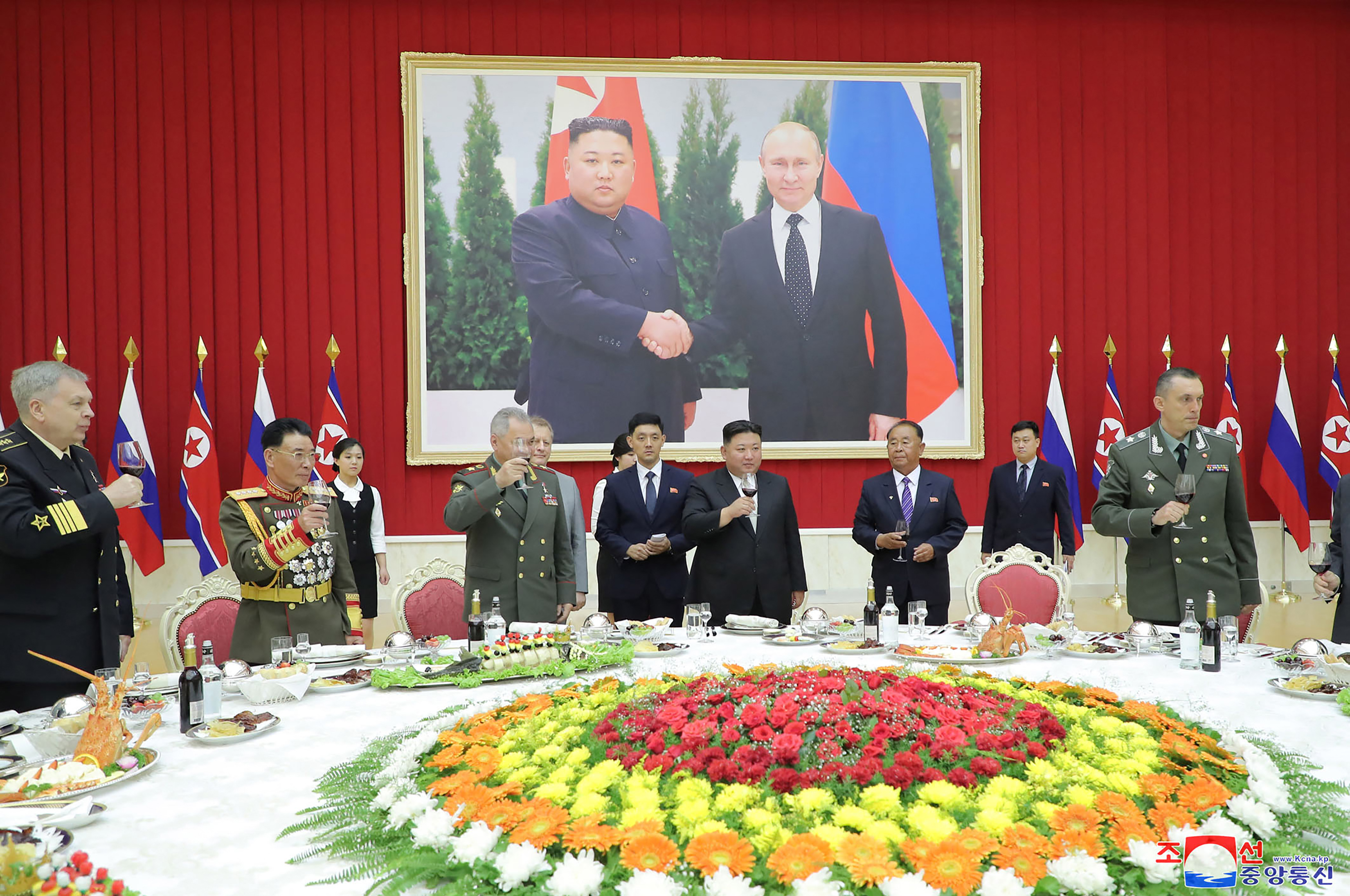 Kim Jong Un and Russian Defence Minister Sergei Shoigu at a banquet. They are making a toast. There is a large display of flowers on the table in front of them, and a large picture of Kim shaking hands with Russian President Vladimir Putin on the wall behind them.