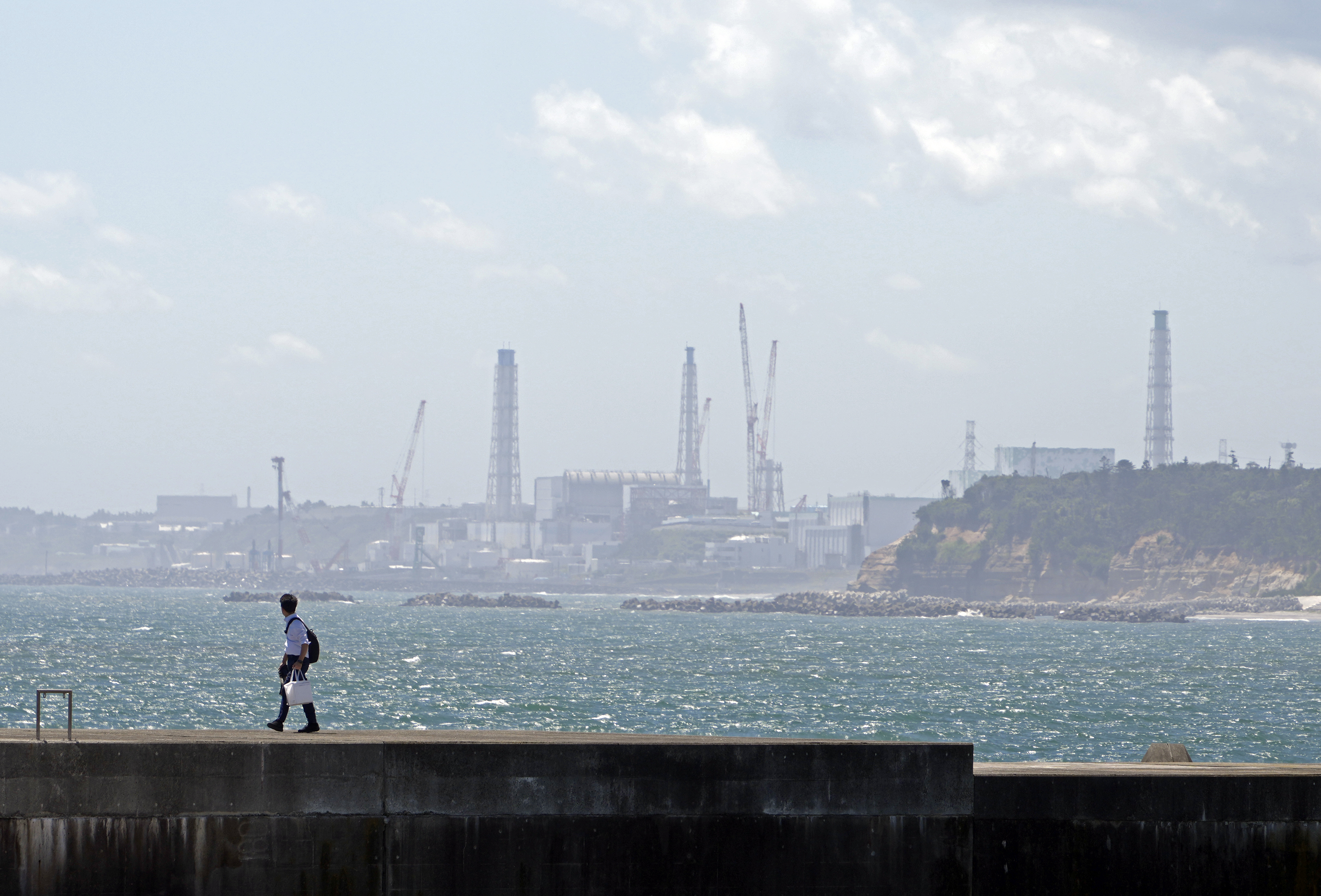 A view of the Fukushima nuclear plant from Namie. The sea is blue and sparkling. Someone is walking on a breakwater.