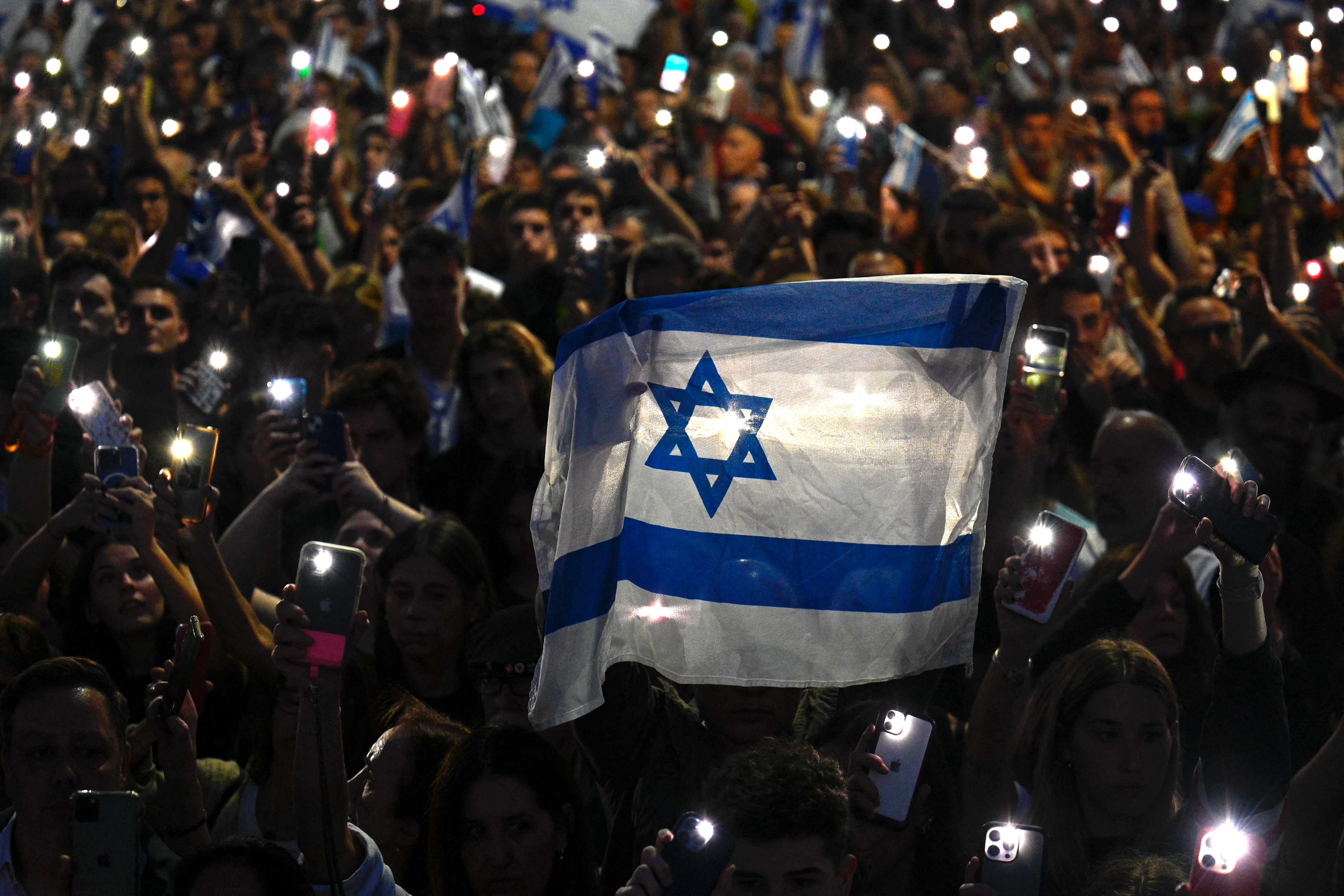 In Israeli flag is held in the foreground as people raise their hands holding glowing lights from their phones in a demonstration at night