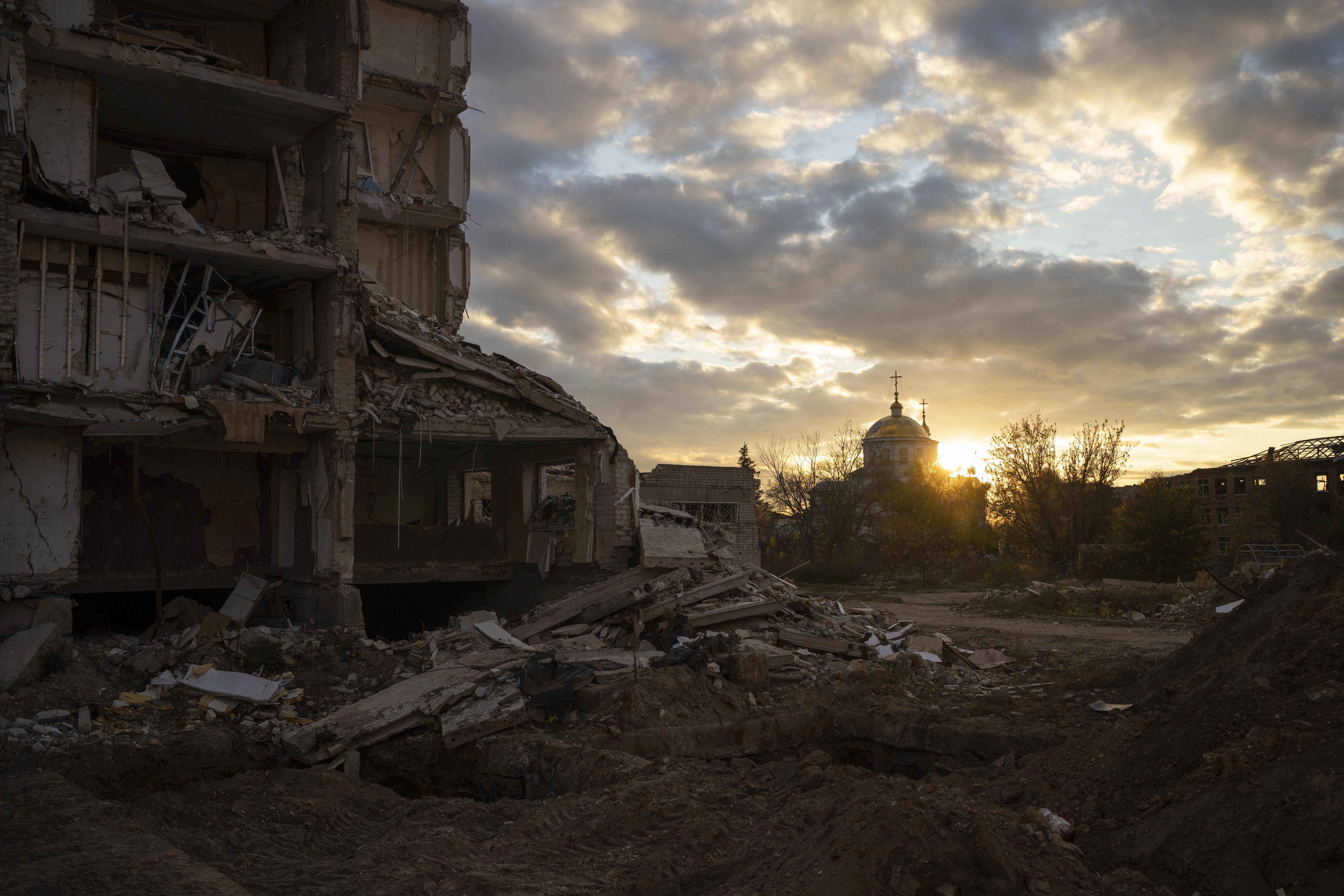 The sun setting over a destroyed building in Izyum. The clouds are tinged in yellow. There s rubble on the ground.