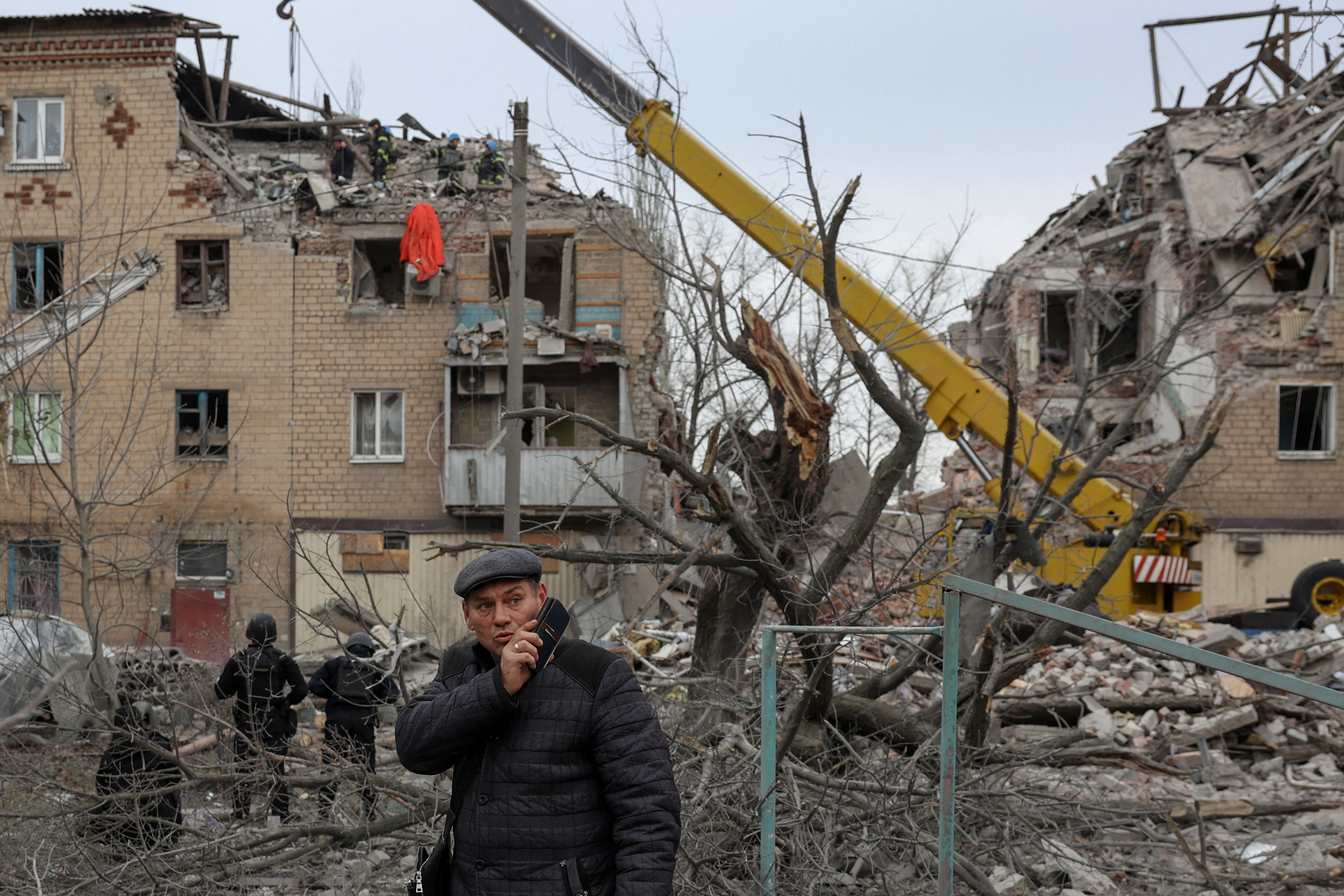 A badly damaged apartment block, with piles of rubble on the ground. There is a man in front on a mobile phone, and workers and a crane behind him.