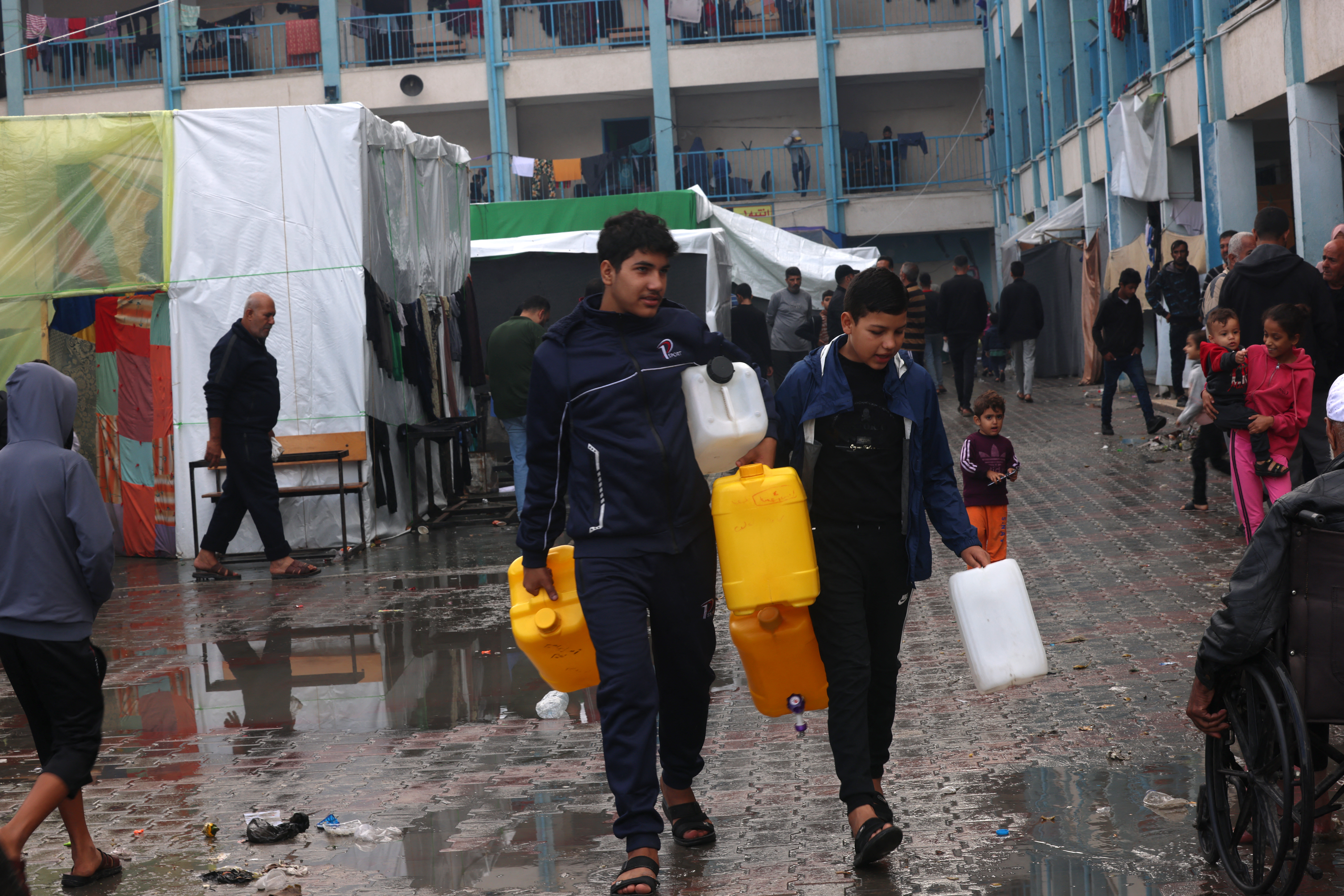 Internally displaced Palestinian boys living in a school run by the UN agency for Palestinian refugees (UNRWA), carry empty water containers as they walk across a wet courtyard following overnight rainstorms in Rafah in the southern Gaza Strip on November 15, 2023 [SAID KHATIB / AFP]