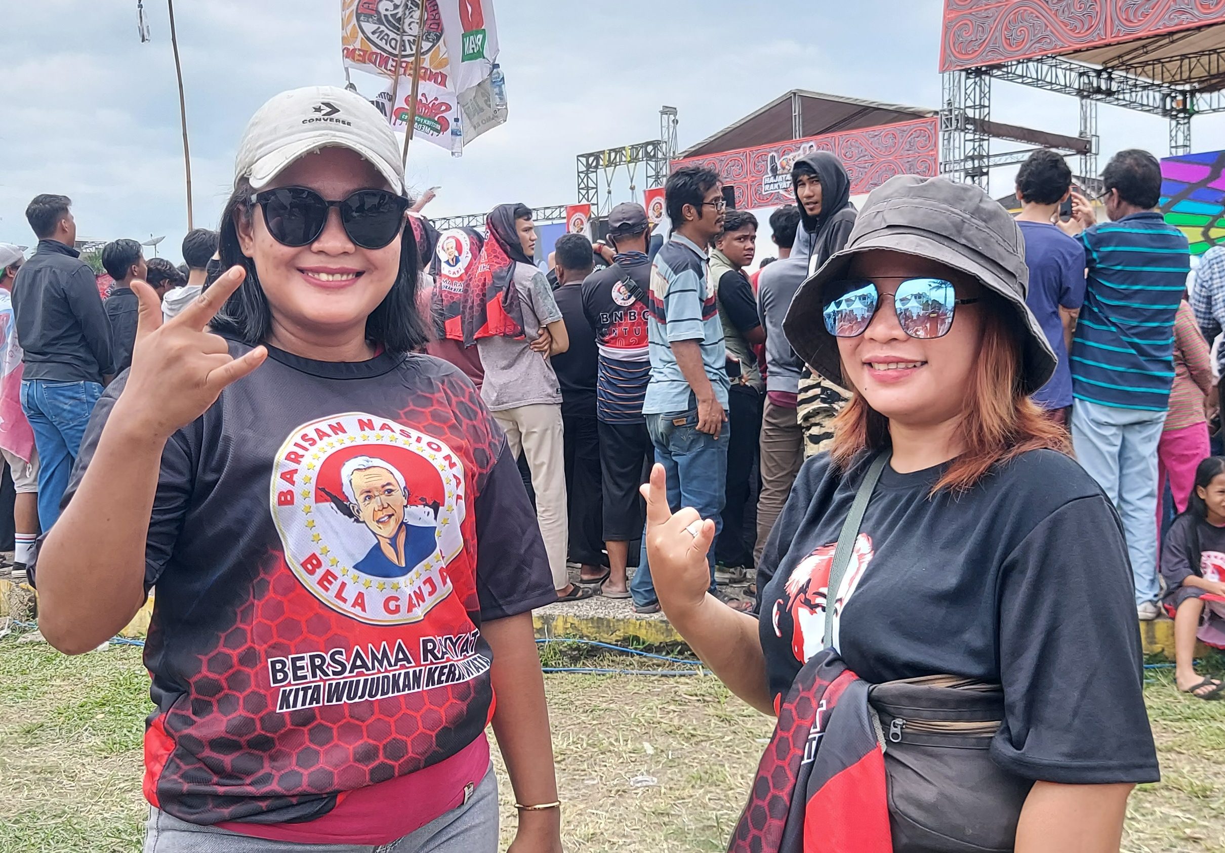 Market sellers Ratna (left) and Lisa (right) are fans of Slank and support Ganjar. Ranta is wearing a T-shirt with Ganjar's face. The stage is behind them. Some people are waving flags.