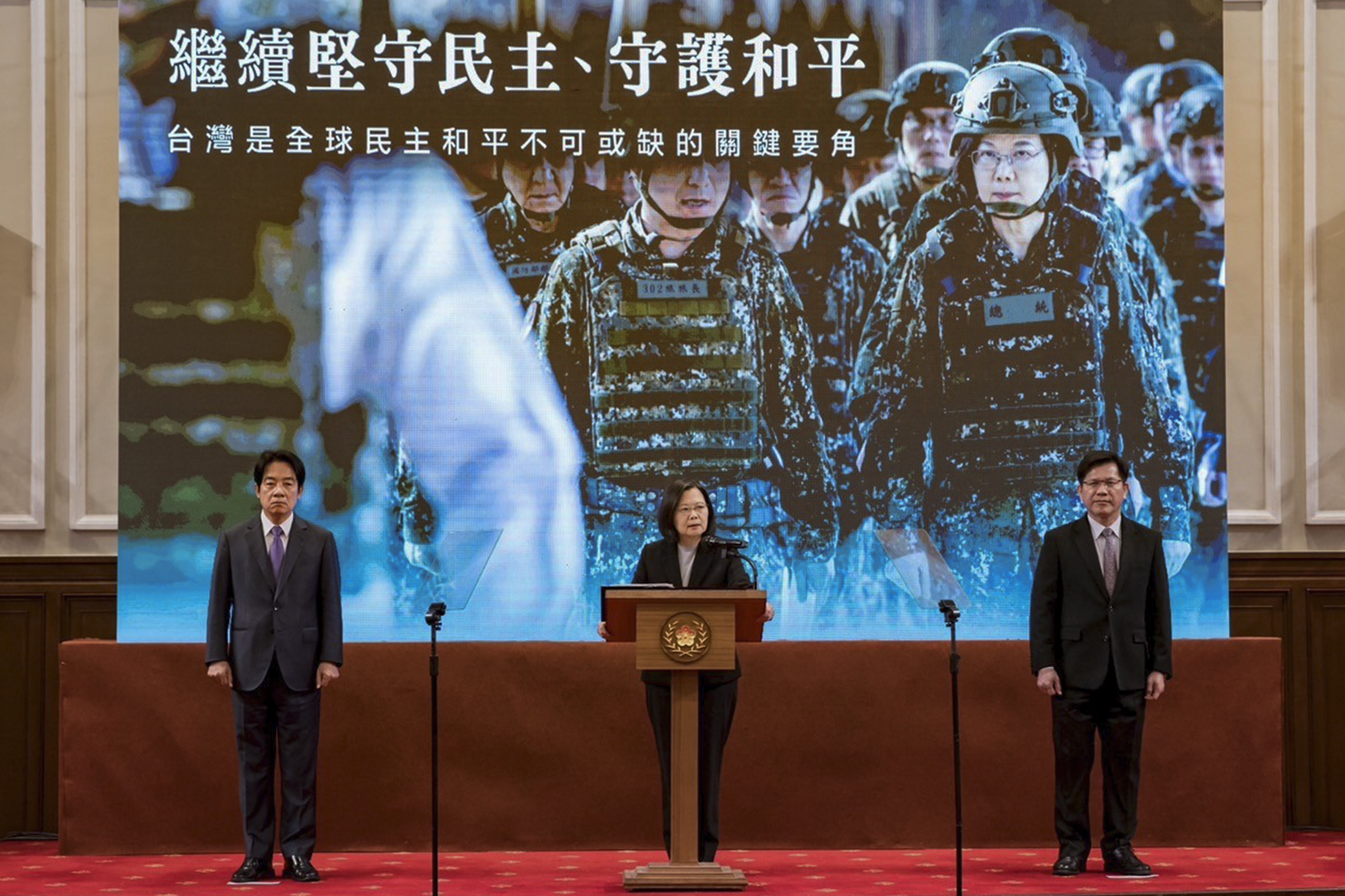 Tsai Ing-wen delivering her New Year speech. She is standing at a lectern with Vice President William Lai on one side. There is a big poster behind showing Tsai in fatigues with the military. The Chinese characters read 'Continue to uphold democracy and safeguard peace, Taiwan is an indispensable key player for global democratic peace'