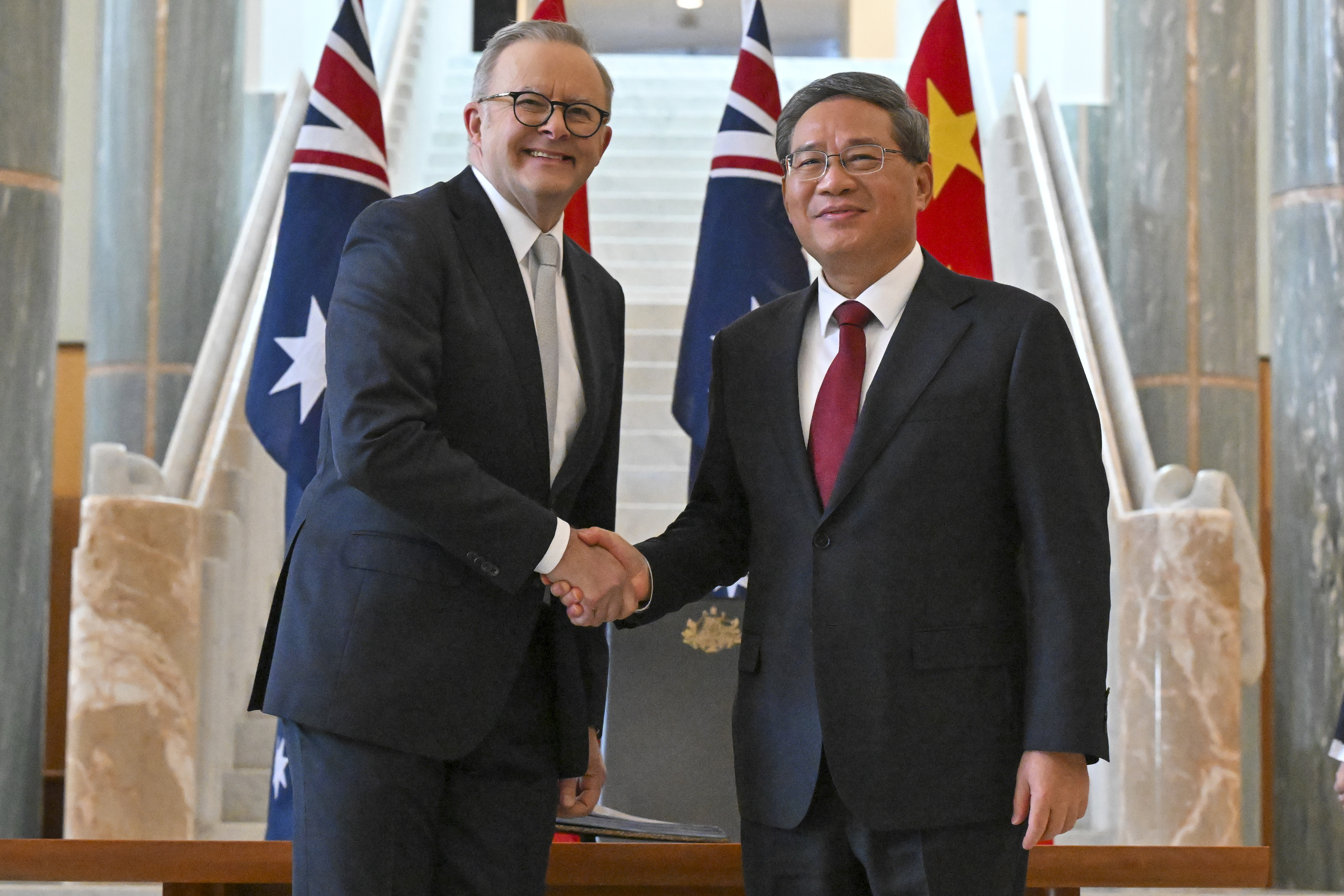 Anthony Albanese and Li Qiang shaking hands. They are smiling. Their country's flags are behind them.