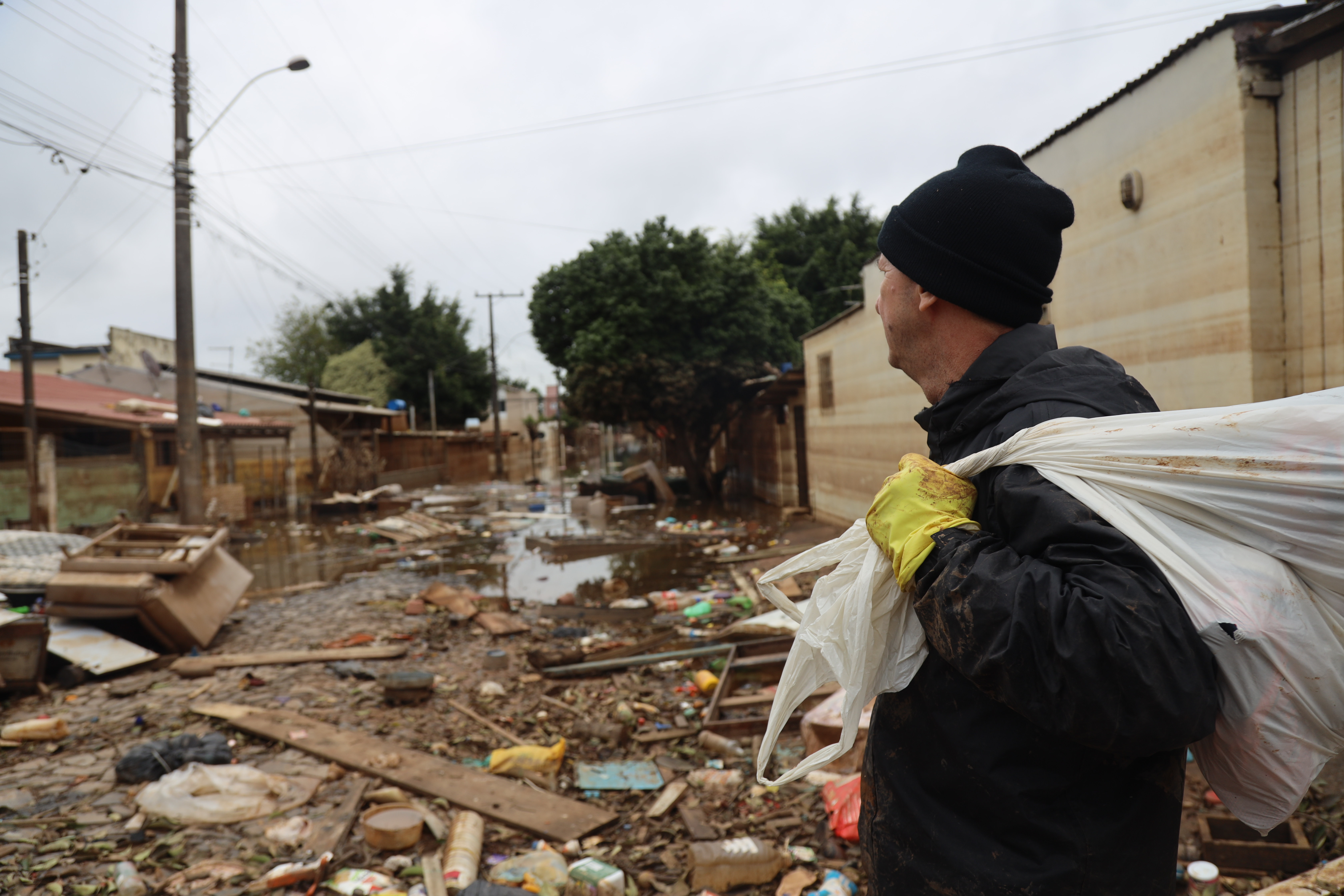 A man with a trash bag slung over his shoulder looks at a neighborhood devastated by floods, the ground still muddy and loose boards and detritus strewn through the streets.