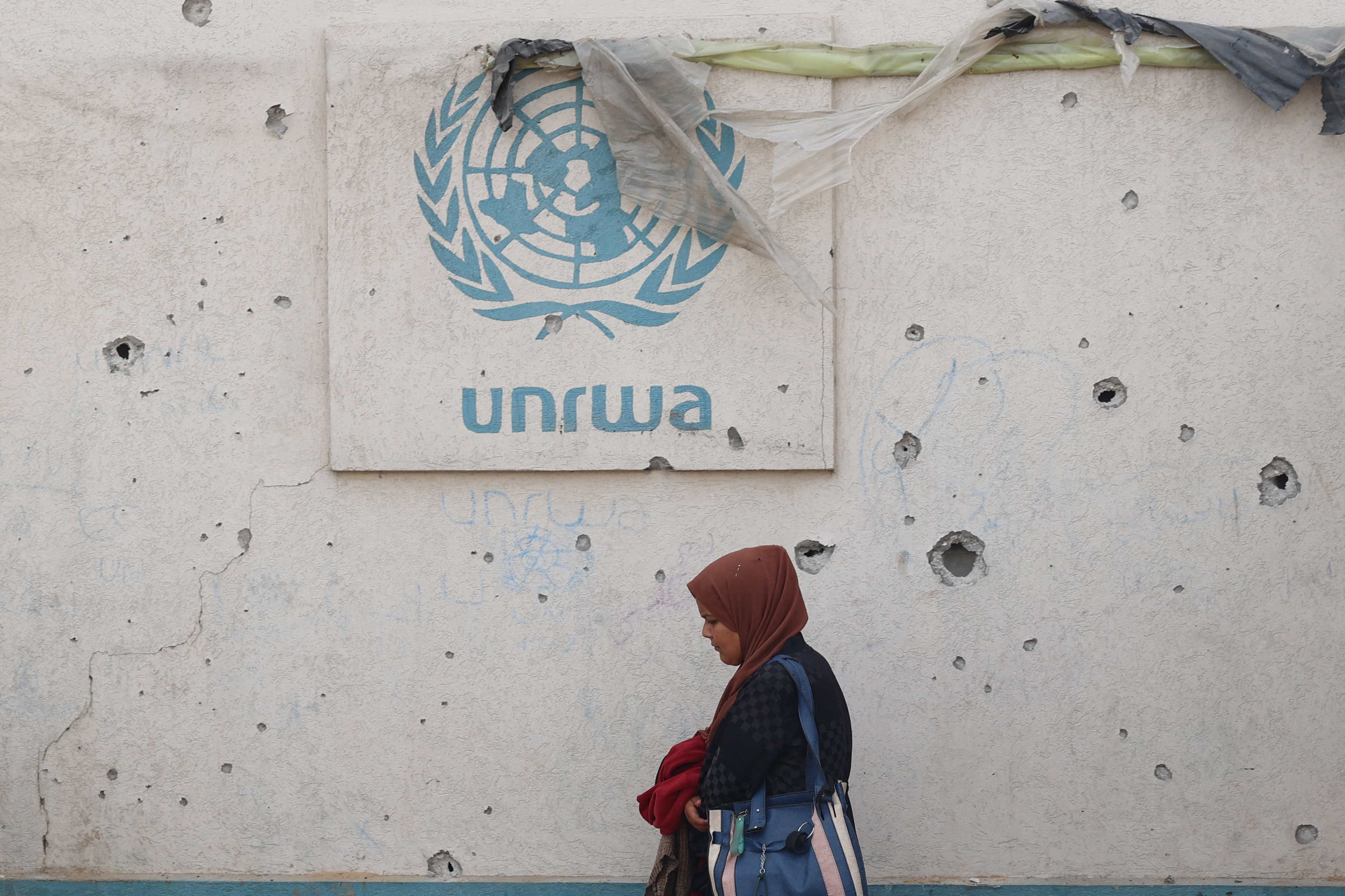 A Palestinian woman walks past a damaged wall bearing the UNRWA logo at a camp for internally displaced people in Rafah in the southern Gaza Strip on May 28, 2024, amid the ongoing conflict between Israel and the Palestinian Hamas militant group. (Photo by Eyad BABA / AFP)