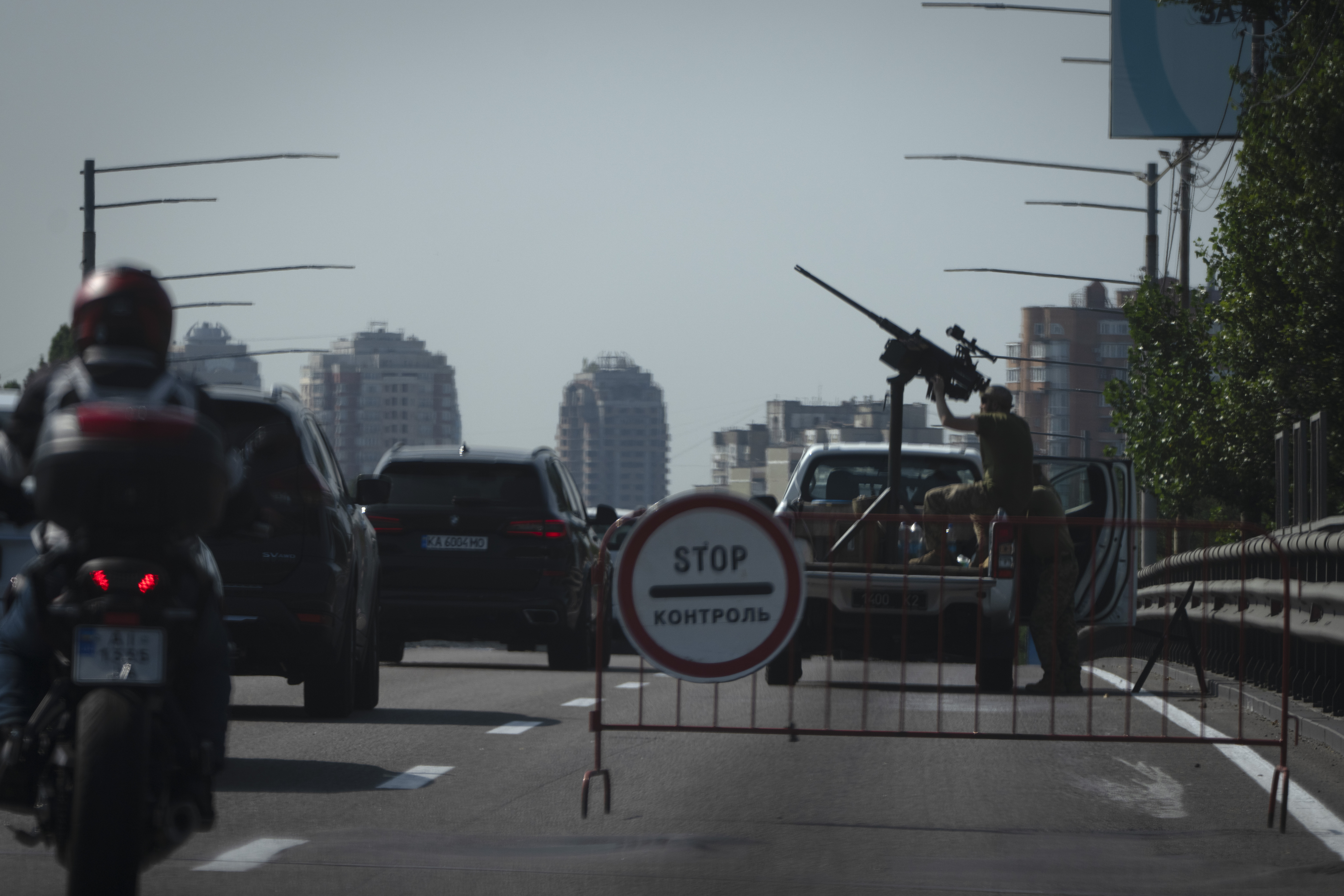 A Ukrainian soldier pointing a weapon towards the sky. The gun is on the back of a truck on the side of the street. There is lots of traffic going past and high rise buildings in the distance