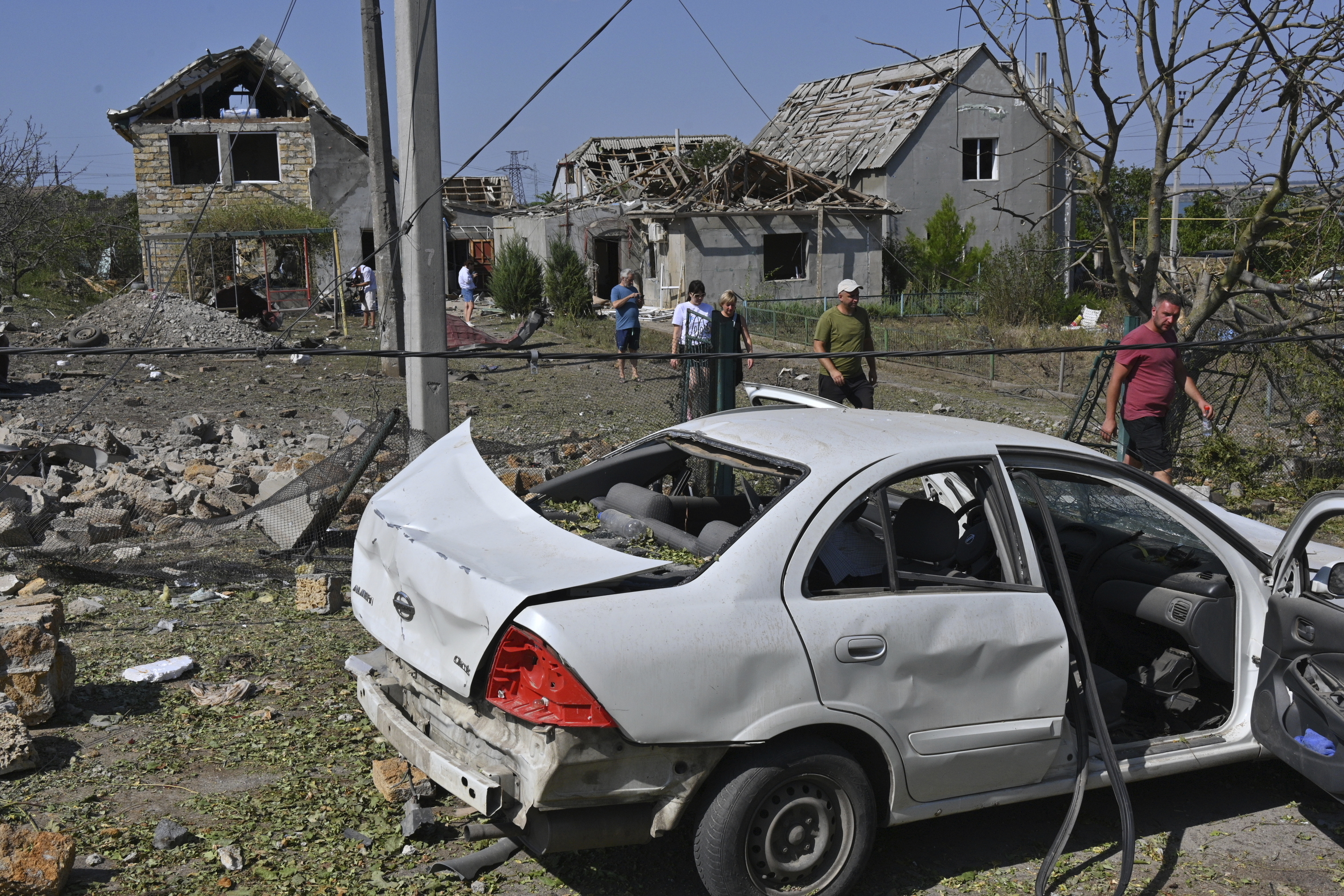 A badly damaged car amid rubble and debris. People are standing around. There are damaged houses in the background.