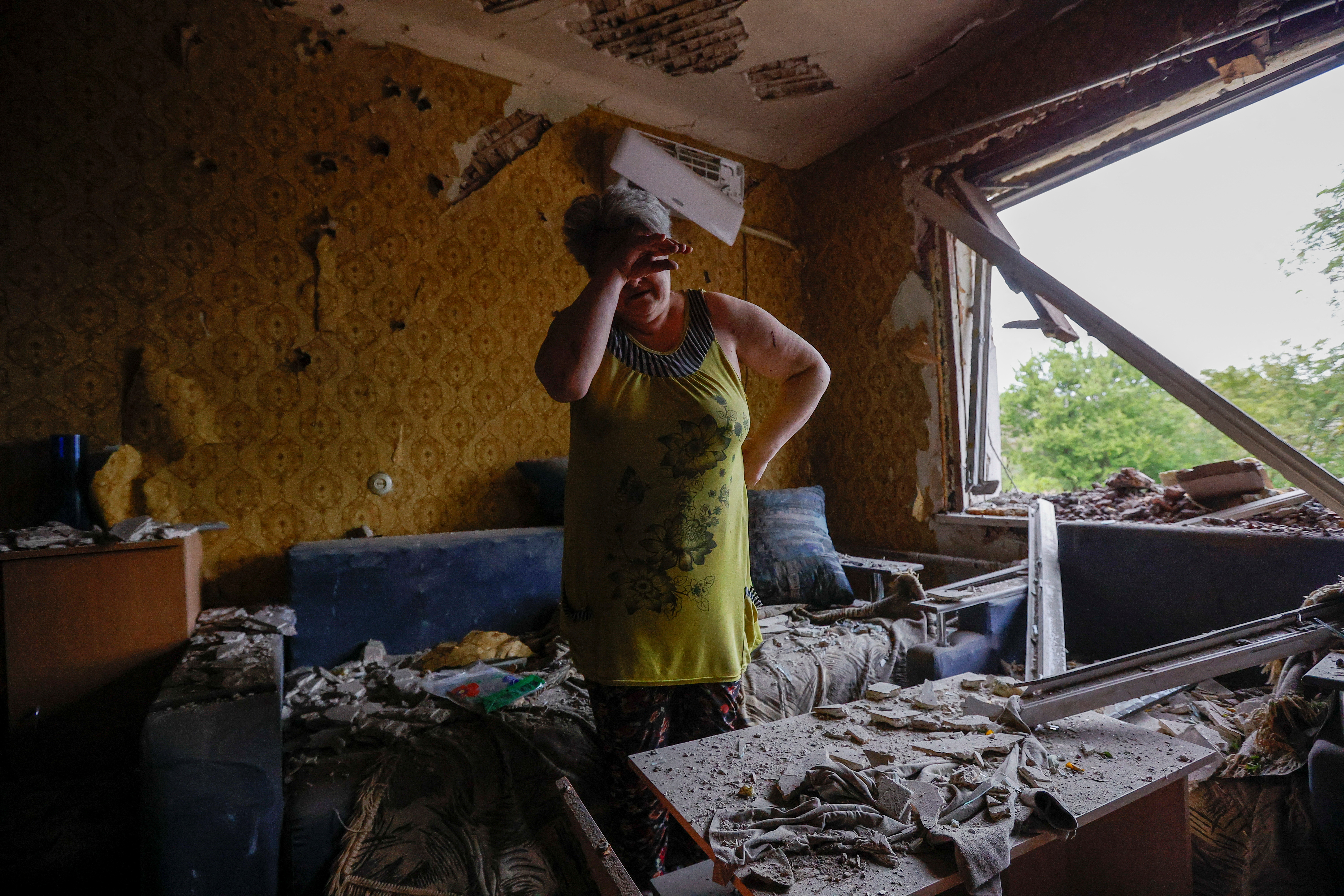 A woman in her ruined apartment. There is no window. The furniture is covered in rubble. She is standing in the middle of the room and has her hand to her face