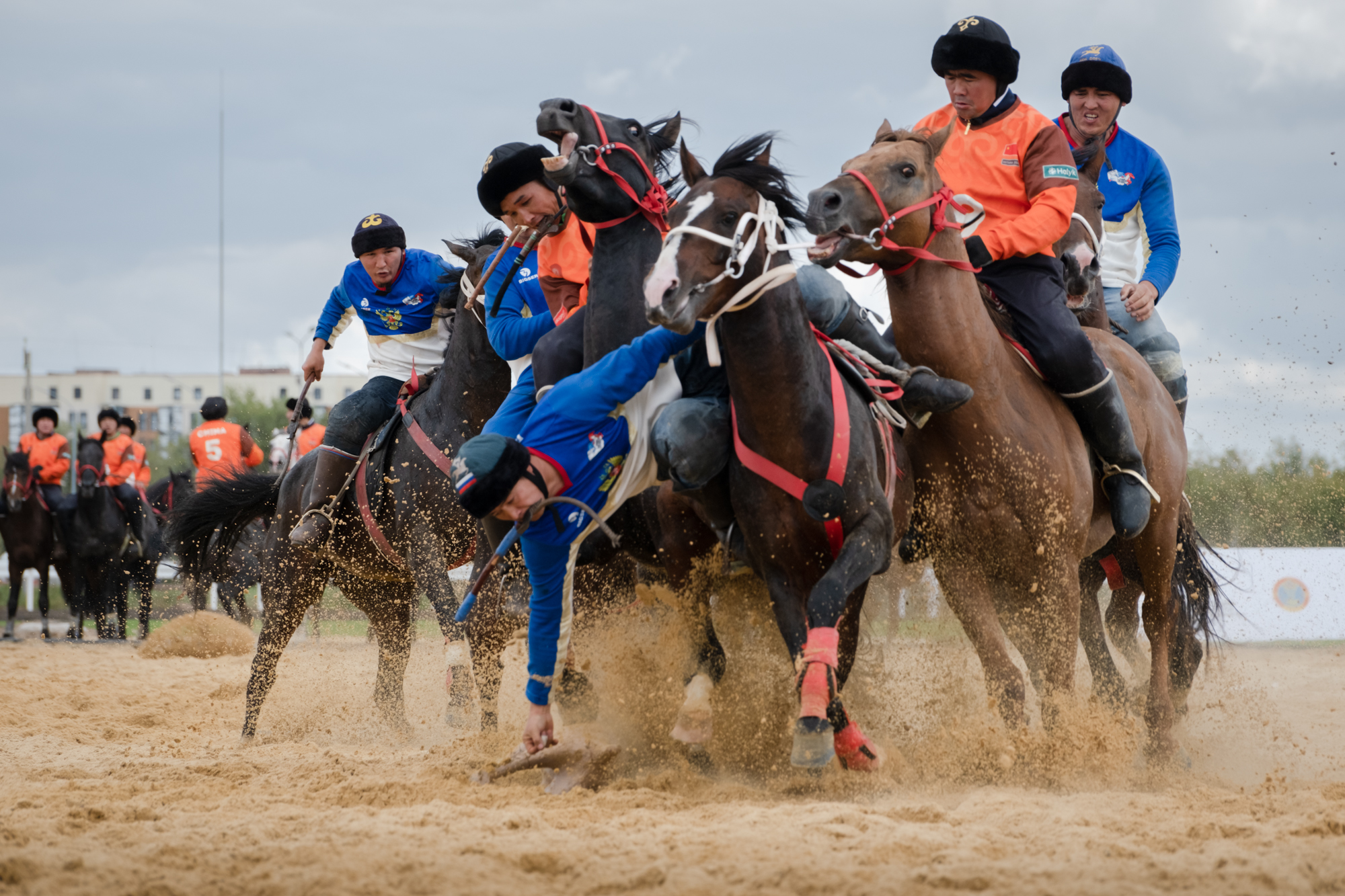 A group of horsemen battling it out in a game of kokpar. There is lots of dist being kicked up. One rider is hanging fro his horse trying to grab something on the ground