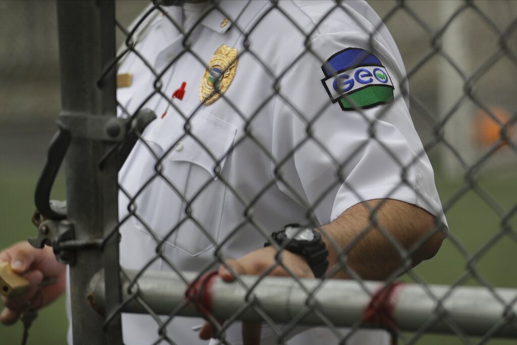 A worker with the GEO Group logo on their shirt behind a chain-link fence