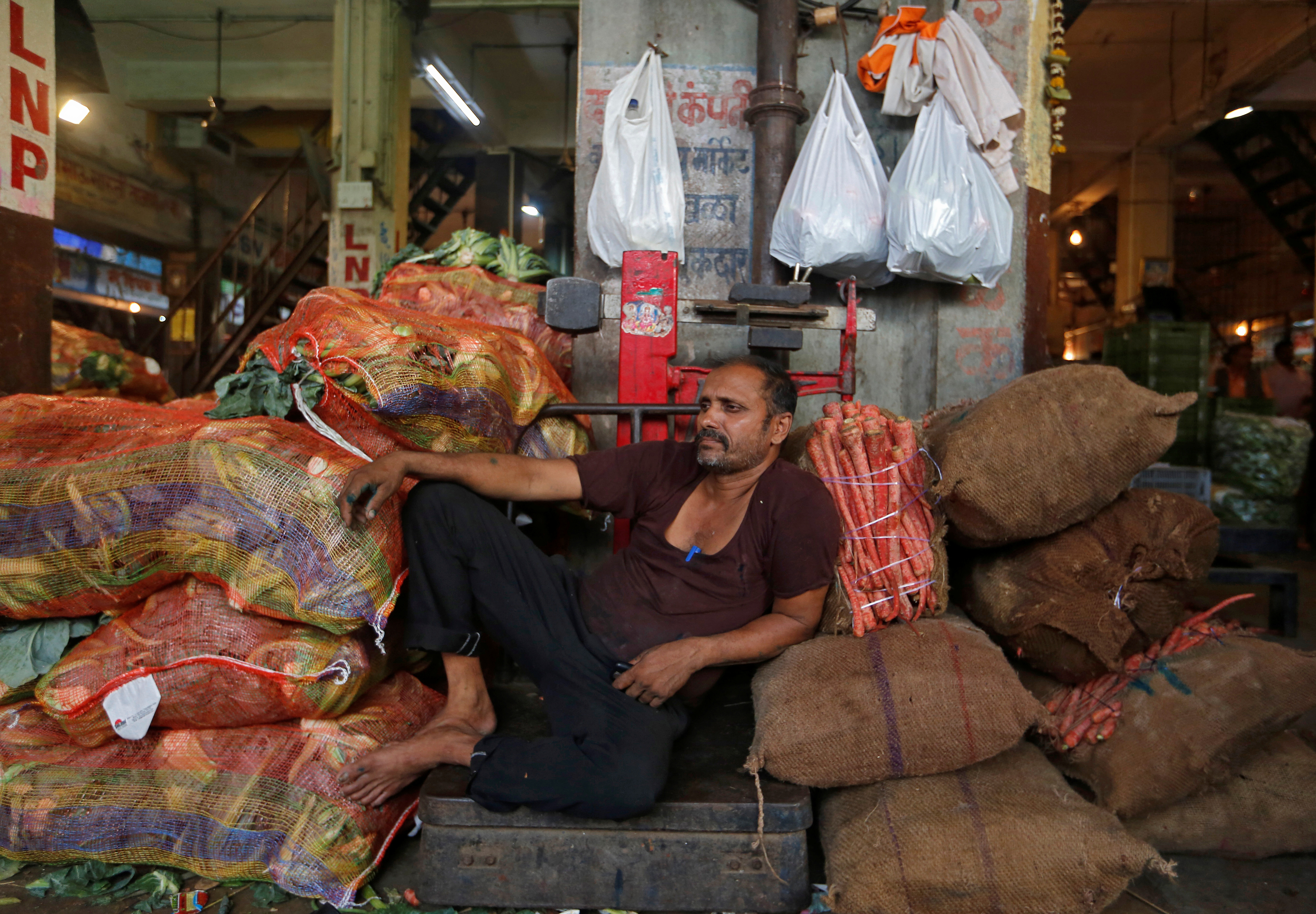 A worker rests on a weighing scale in between the sacks filled with cauliflower and carrots at a wholesale vegetable market in Mumbai, India