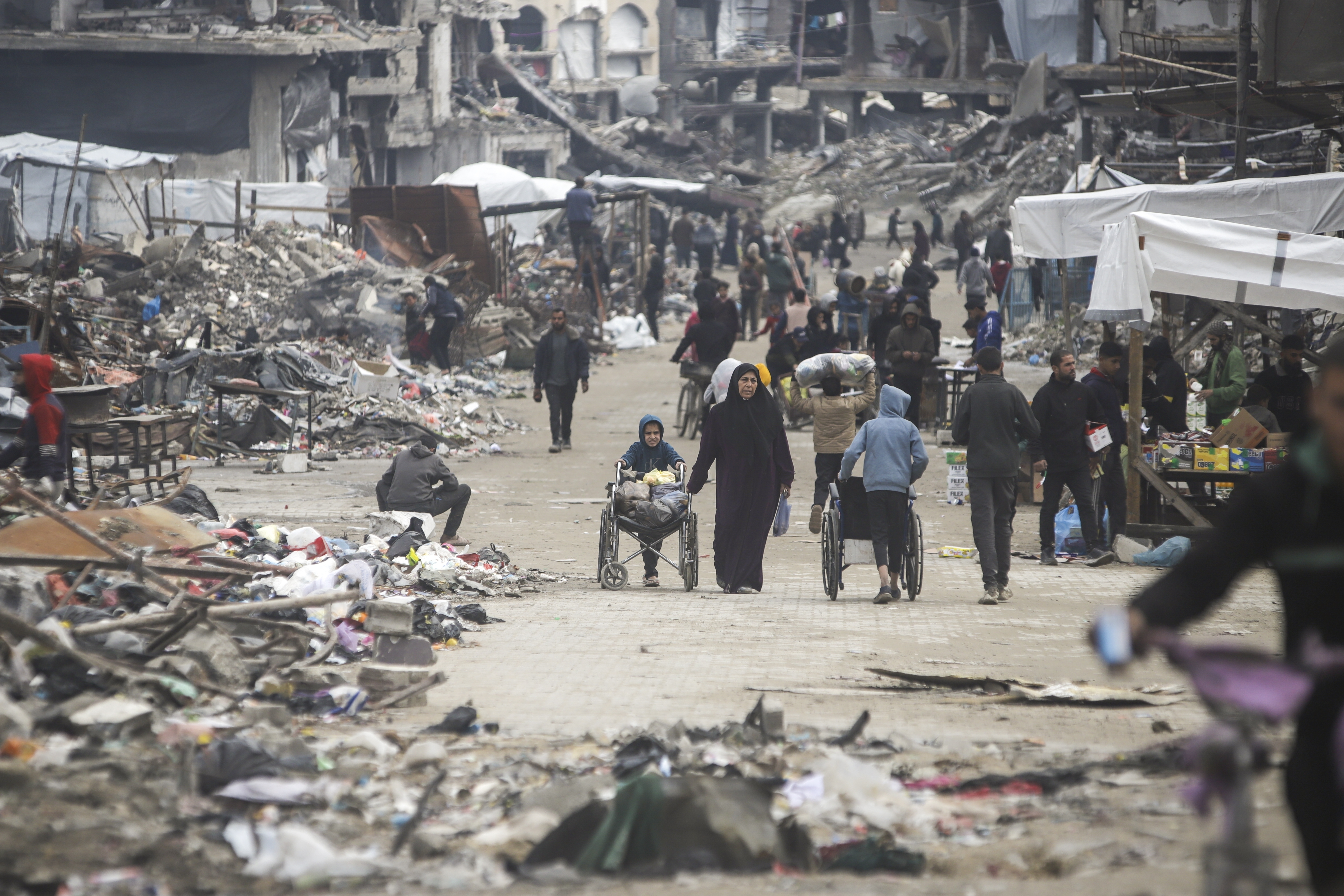 Pedestrians walk along a road lined with few stands selling goods, amid widespread destruction