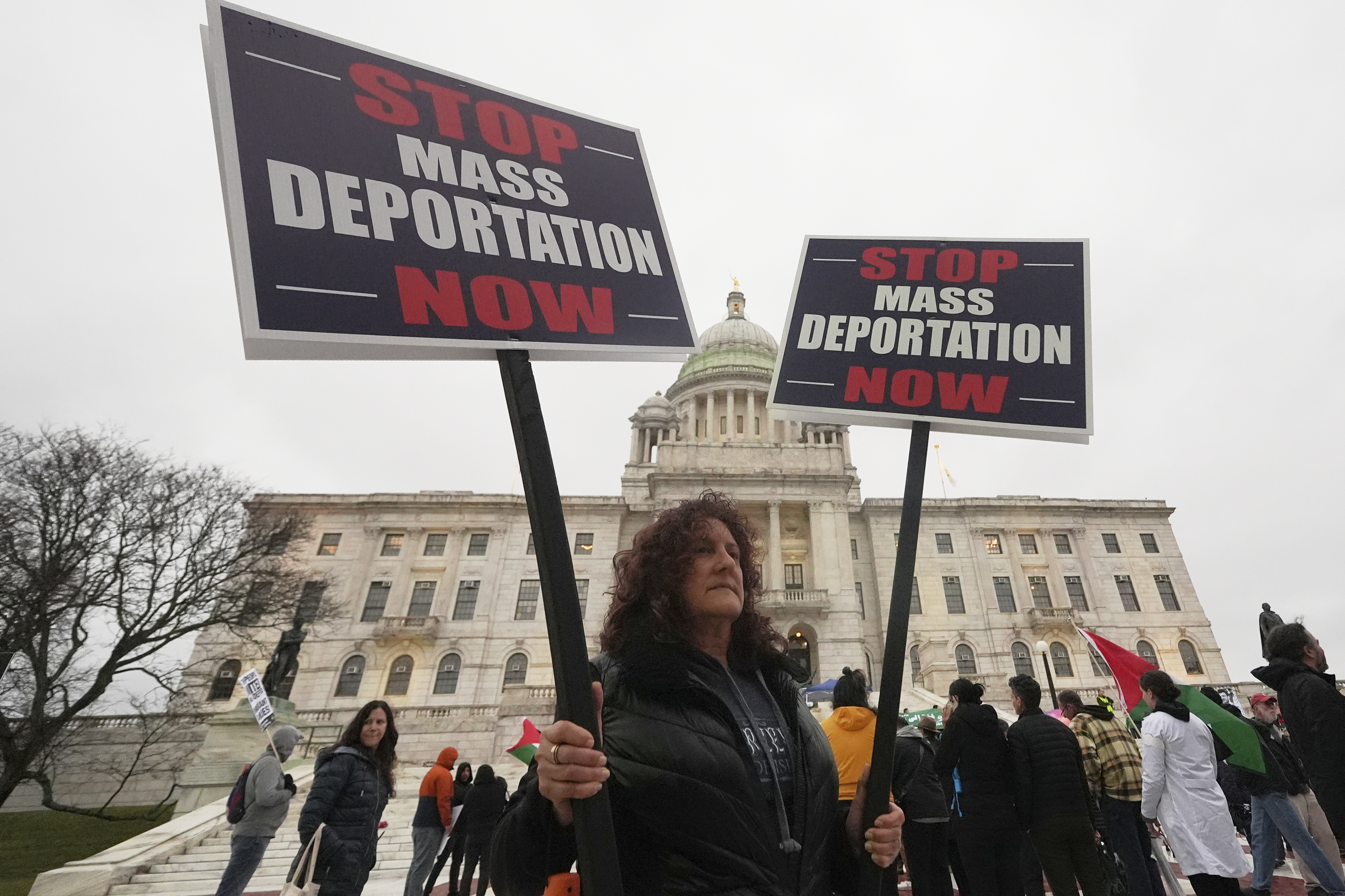 A woman in front of the Rhode Island State House holds up signs that read, "Stop Mass Deportation Now."