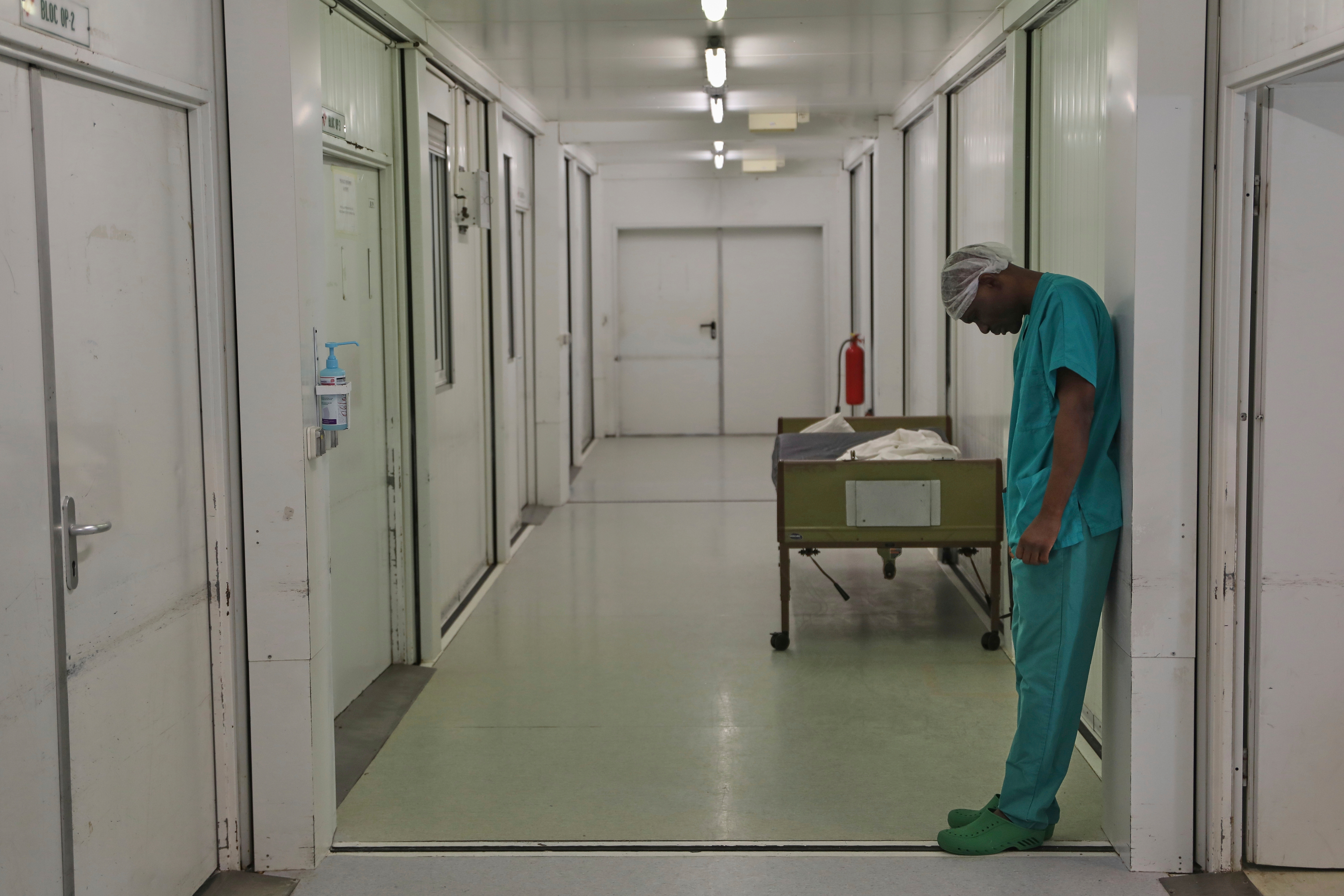 A doctor in an empty hallway leans forward and looks at the ground.