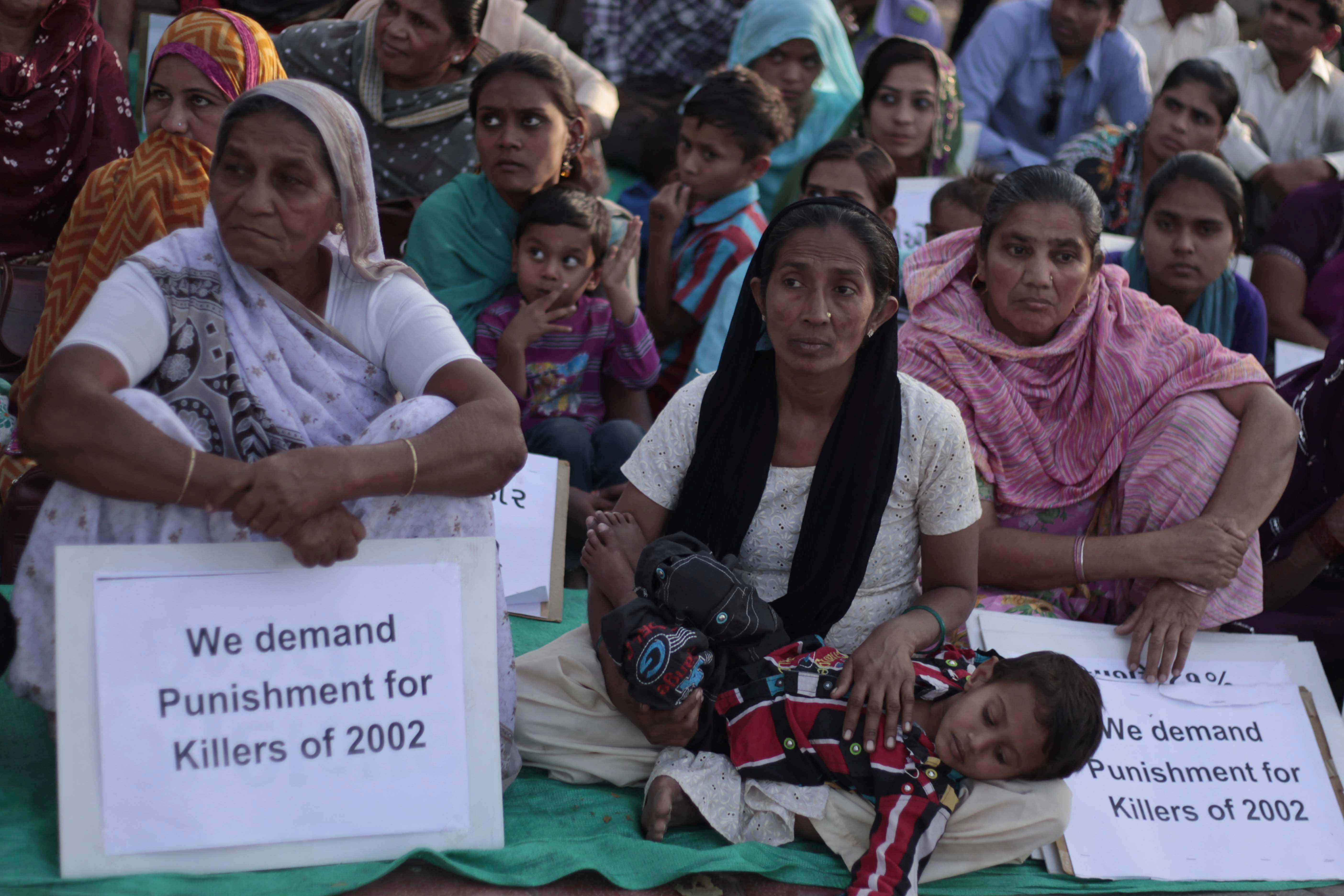 Protesters in Gujarat sit on the ground with protest signs that read "We demand punishment for the killers of 2002"