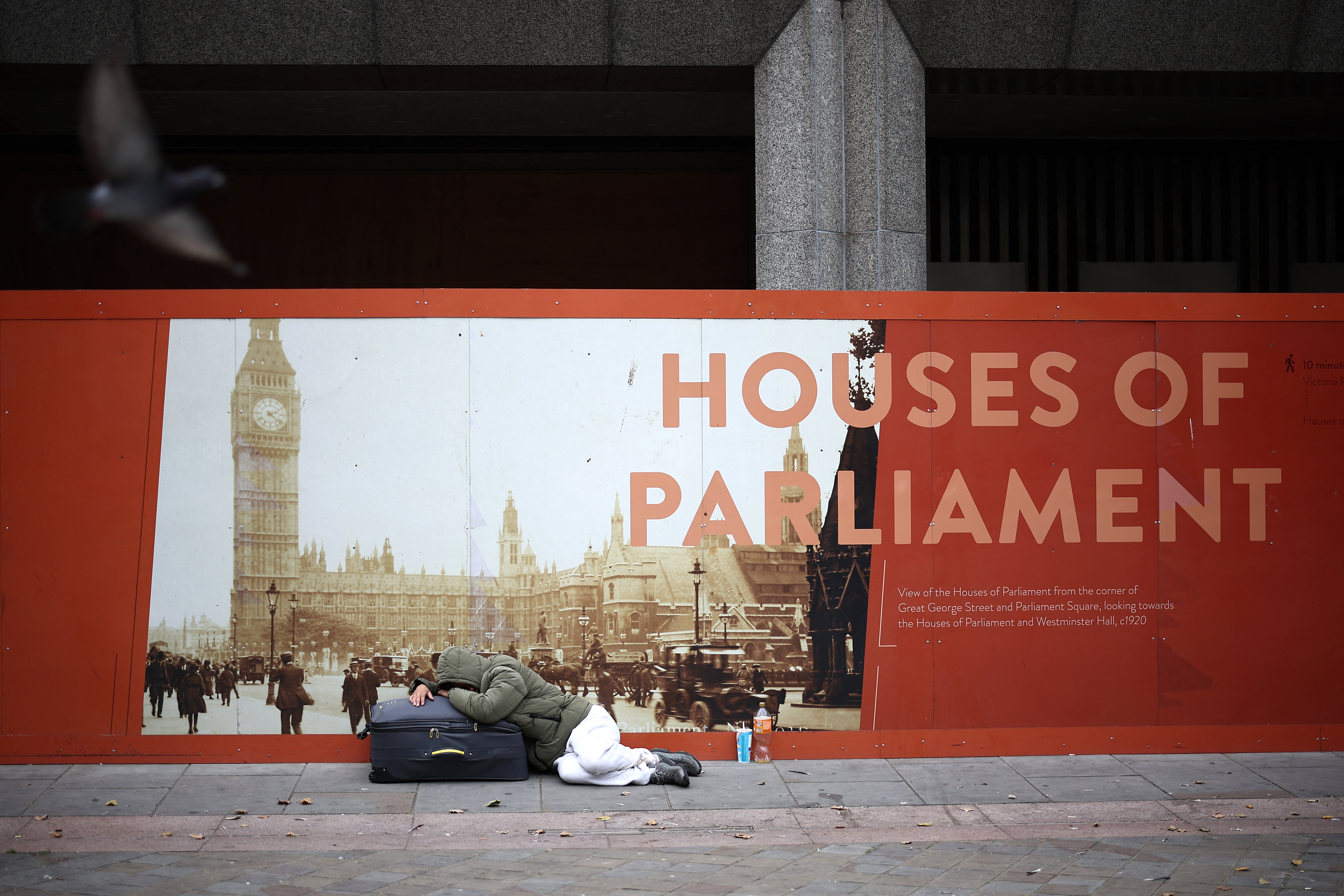 A rough sleeper lies on a suitcase in front of an advertisement board showing the Houses of Parliament in central London on September 15, 2024. (Photo by HENRY NICHOLLS / AFP)