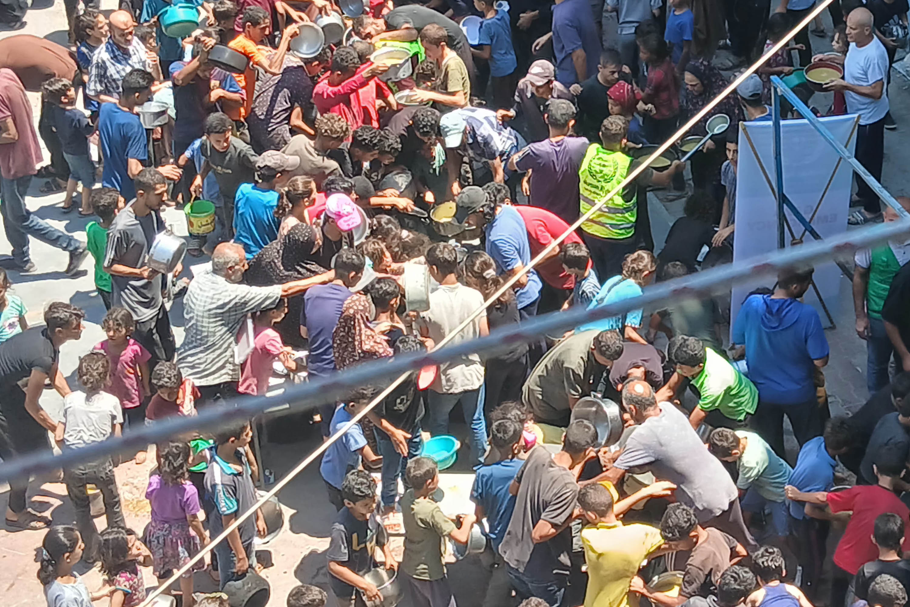 A crowd of people gathered over large containers of cooked lentils in a school yard, seen from a higher floor