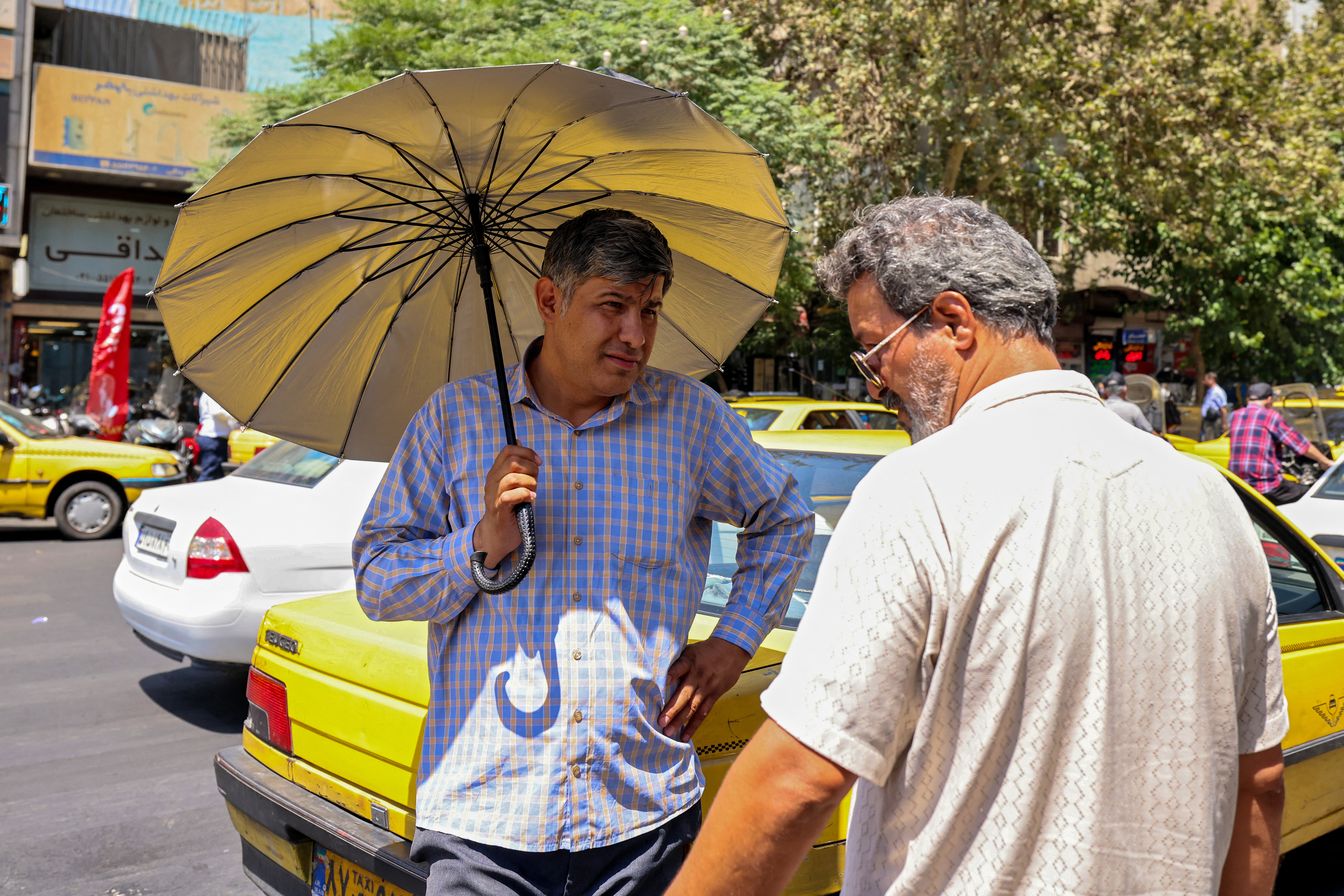Iranians using umbrellas to shield themselves from the sun, chat on a street during a heatwave in the capital Tehran on August 9, 2025. (Photo by ATTA KENARE / AFP)