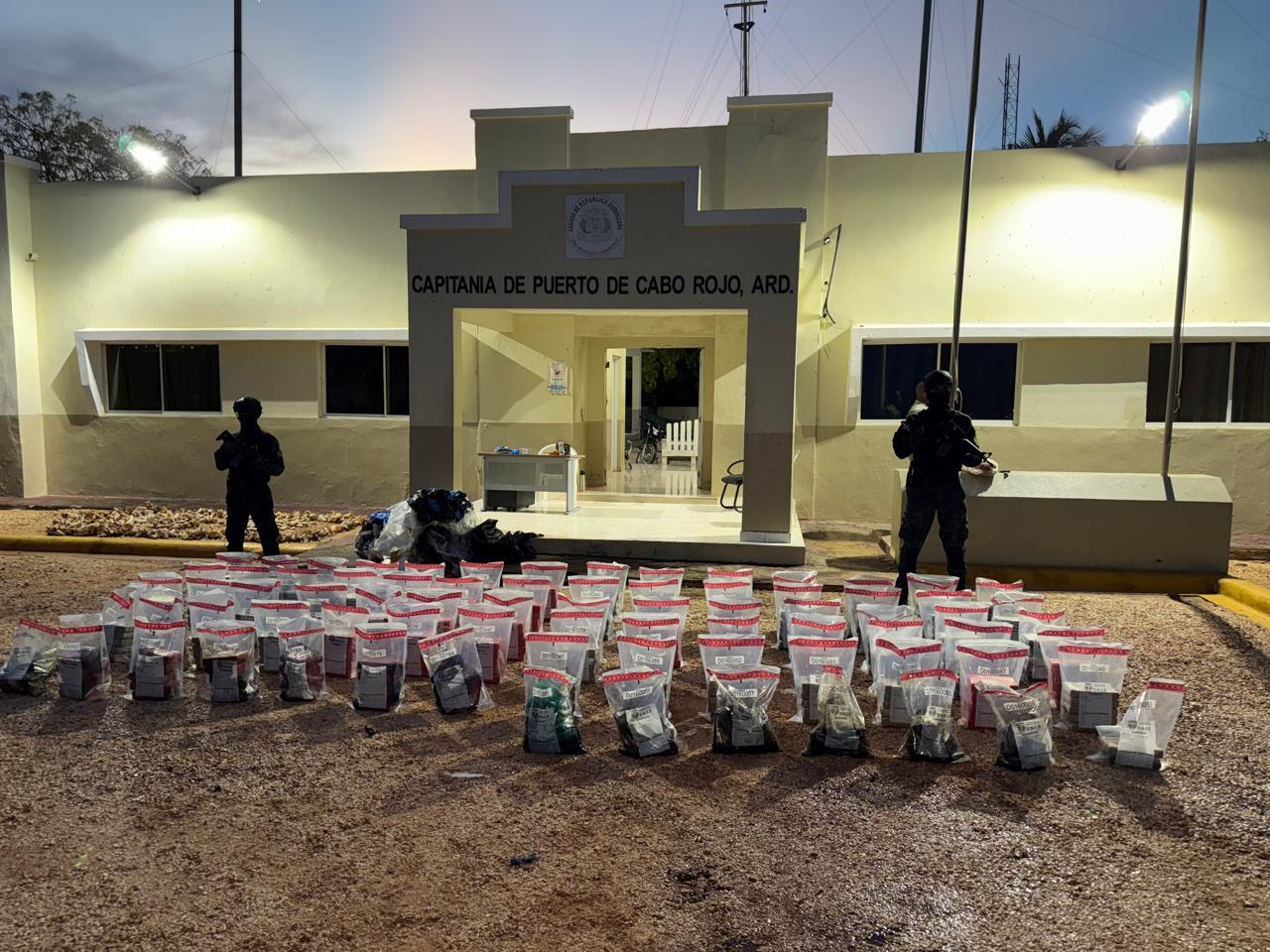 Large number of bags of cocaine lie on the ground in front of armed personnel.