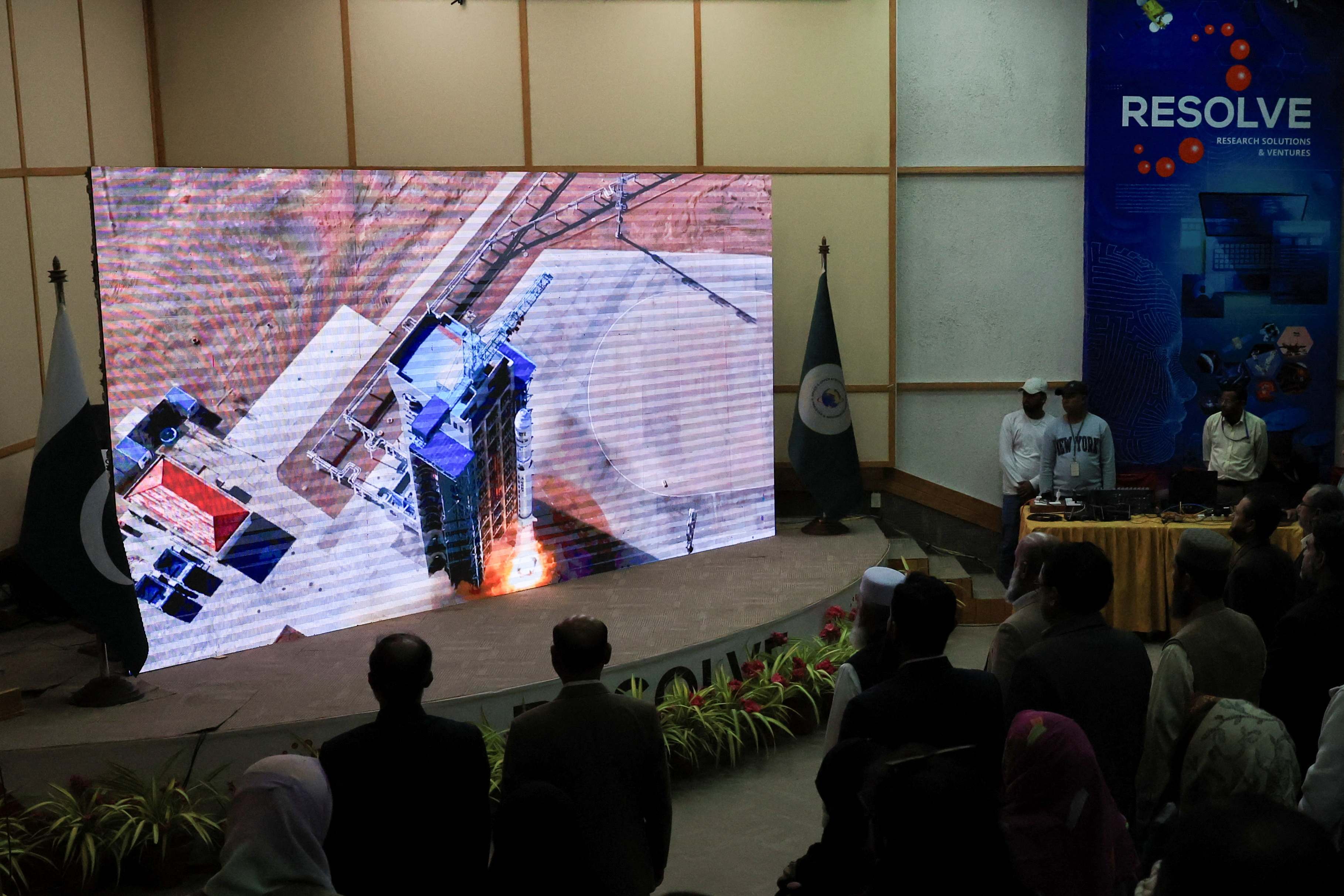 People watch a screen displaying the launch of a satellite.