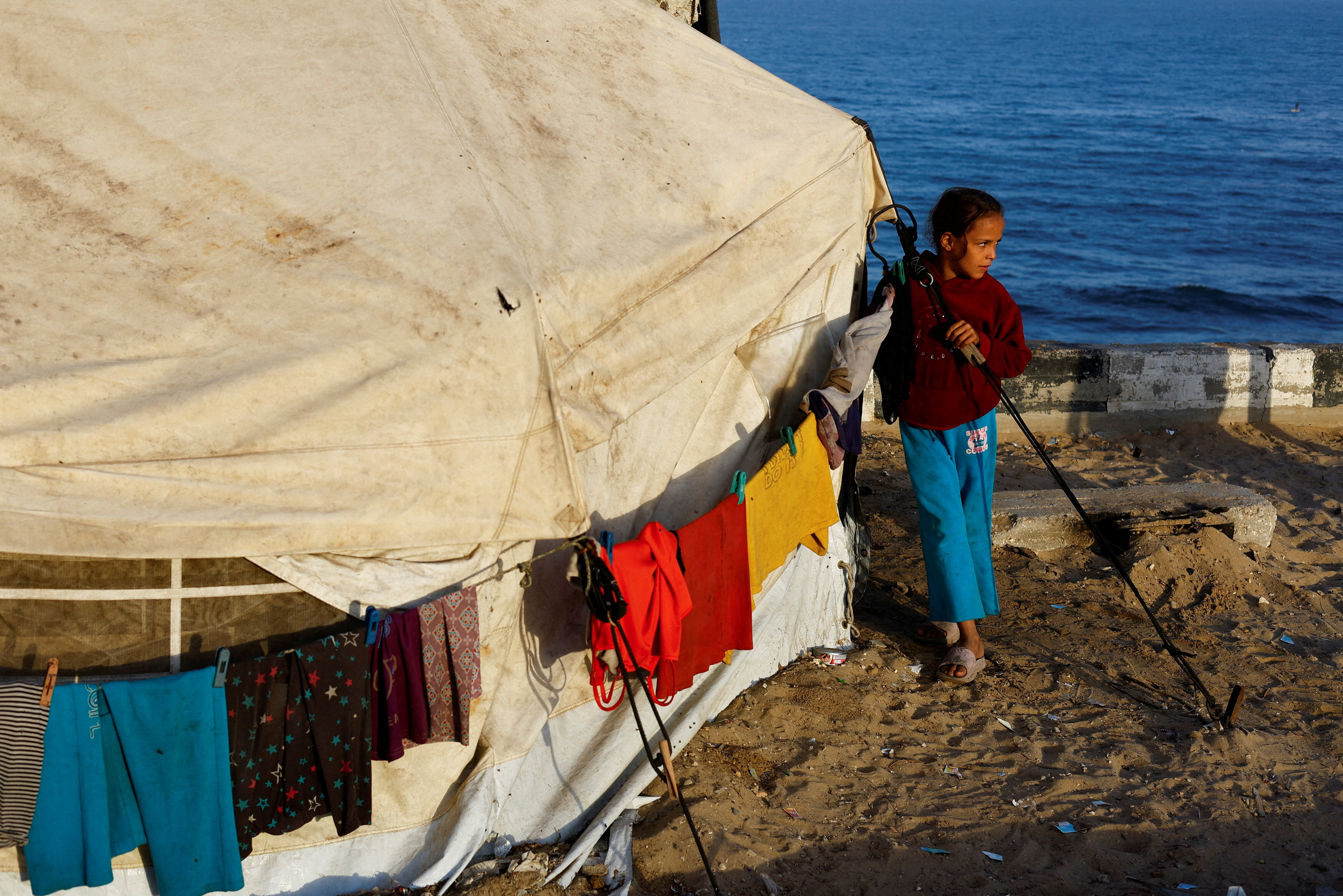 A displaced Palestinian girl stands next to a tent on a roadside.