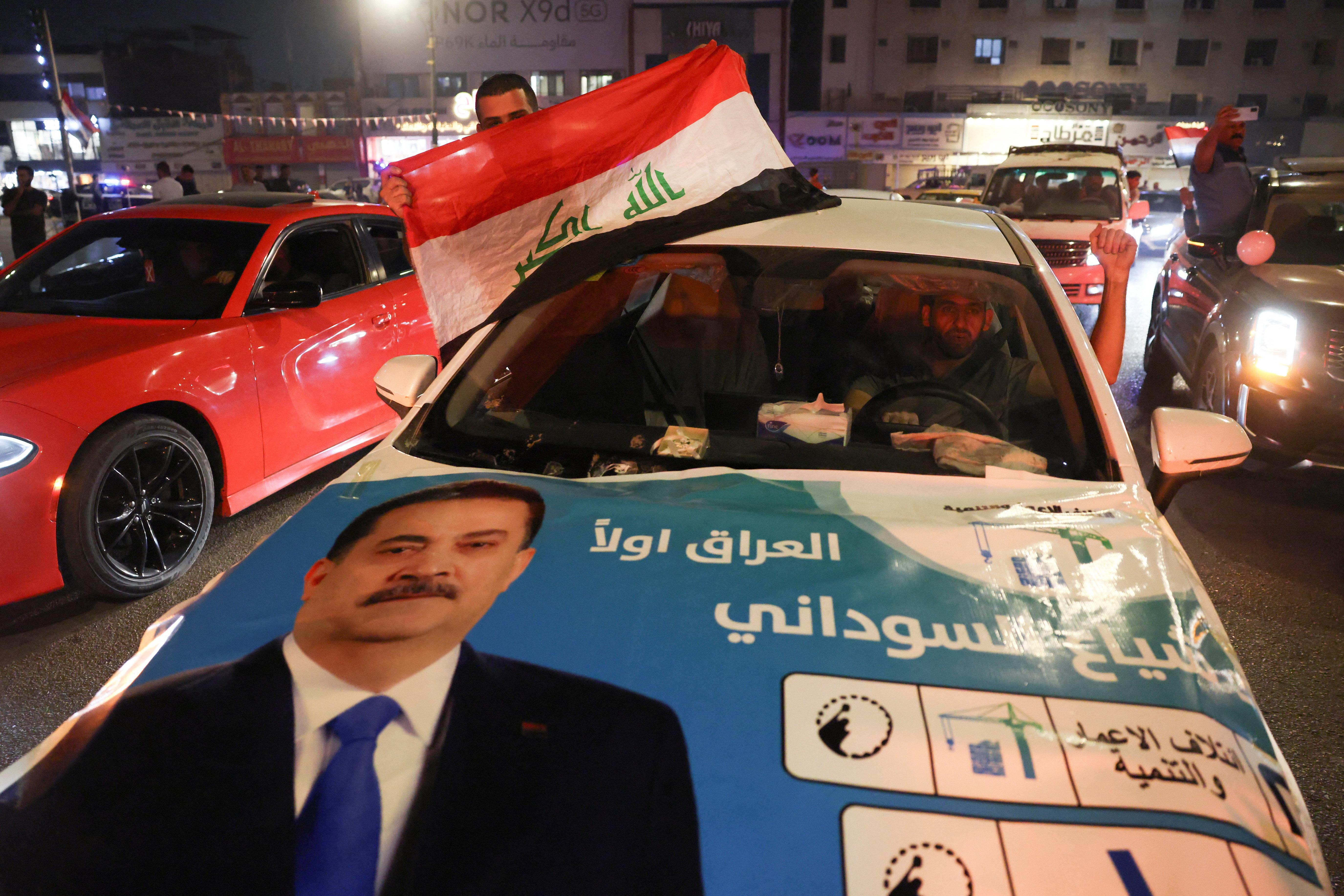 A man holds a flag of Iraq from a car with an election poster featuring the current Prime Minister