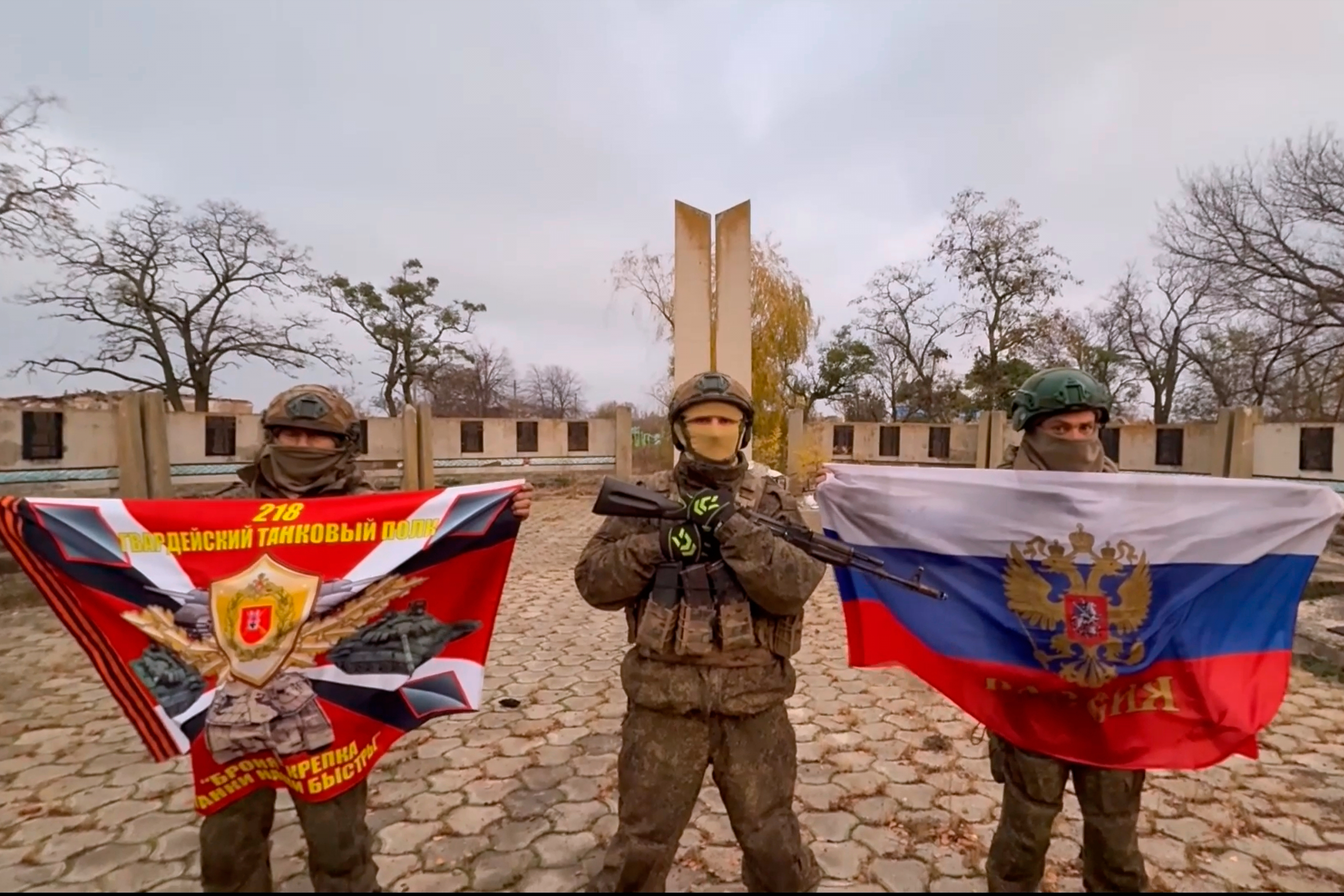 In this image, made from video and provided by Russian Defense Ministry Press Service on Friday, Nov. 7, 2025, Russian soldiers hold a Russian national and their unit flag as they pose for a photo in front of a memorial for soldiers fallen during battle with Nazi German soldiers, after capturing Uspenivka village in Zaporizhzhia region of Ukraine. (Russian Defense Ministry Press Service via AP
