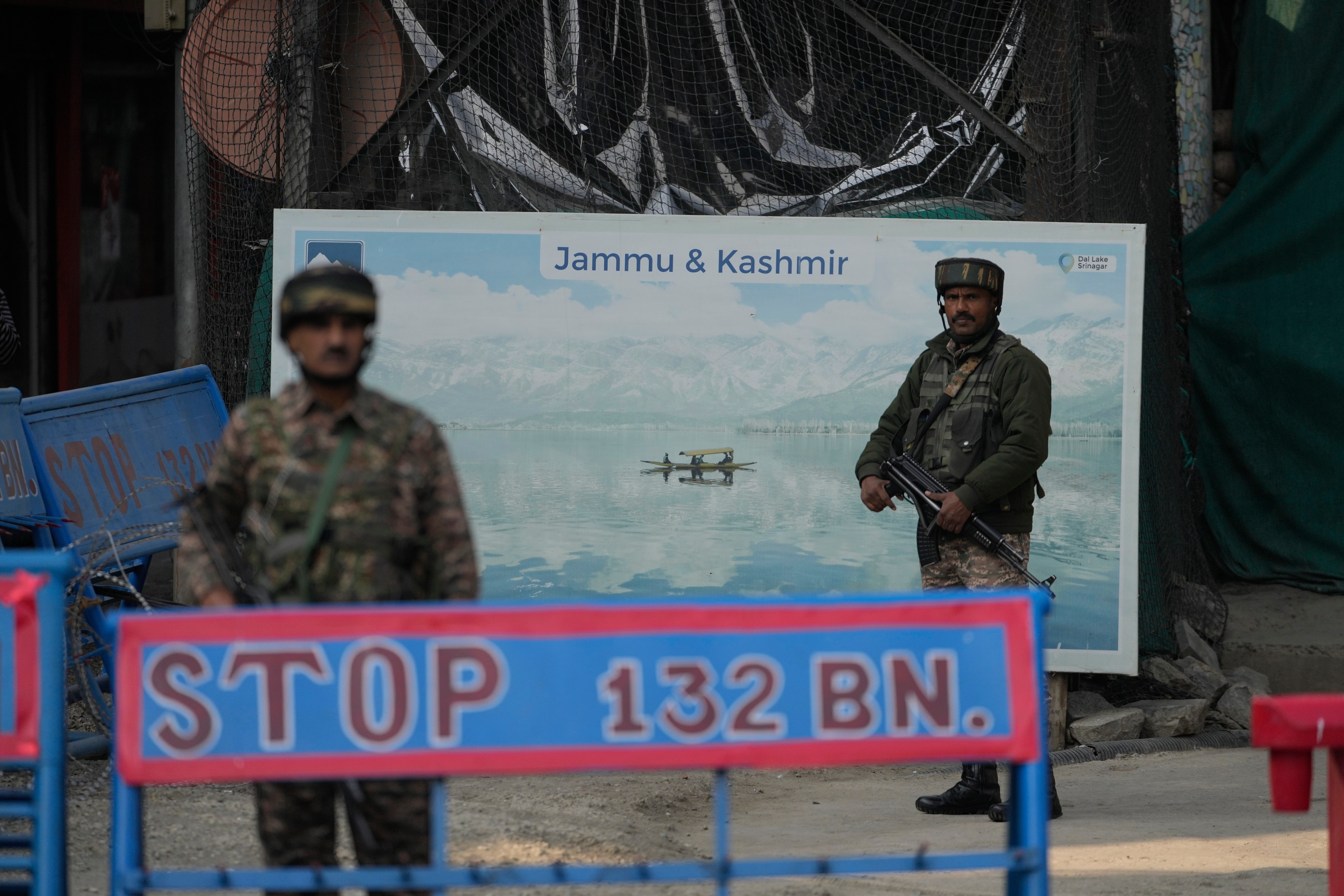 Indian soldiers stand guard at a checkpoint with a poster identifying "Jammu and Kashmir" on the road behind them.